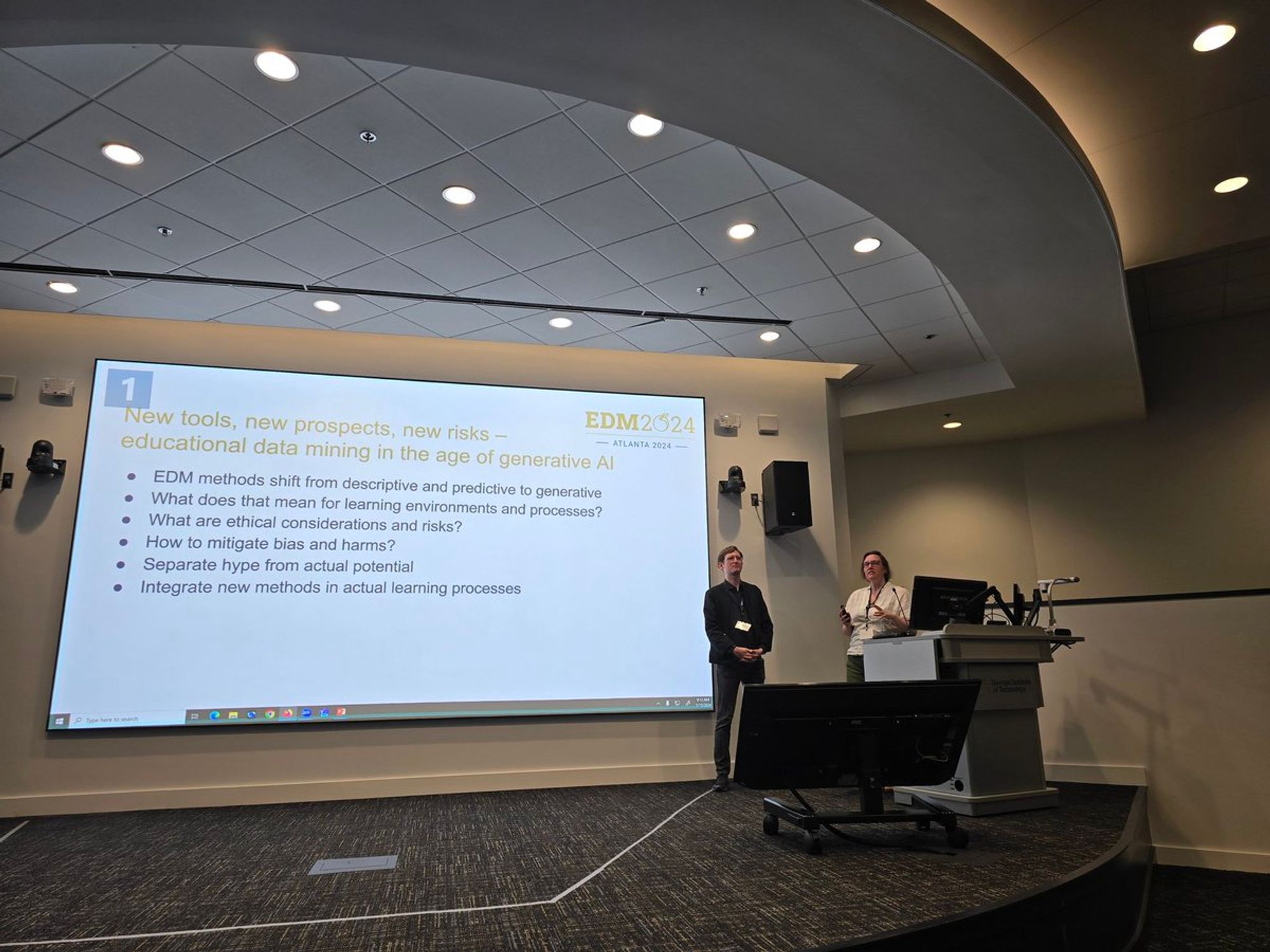 Benjamin Paaßen and Carrie Demmans Epp Chairing the International Conference on Educational Data Mining. Benjamin and Carrie are visible on stage with a slide behind them on the conference motto: New tools, new prospects, new risks - educatinal data mining in the age of generative AI.