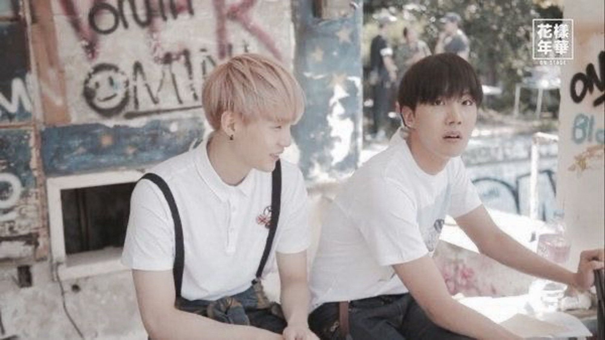 Suga and J-Hope wearing white short sleeved shirts and sitting next to each other in front of graffiti’d walls