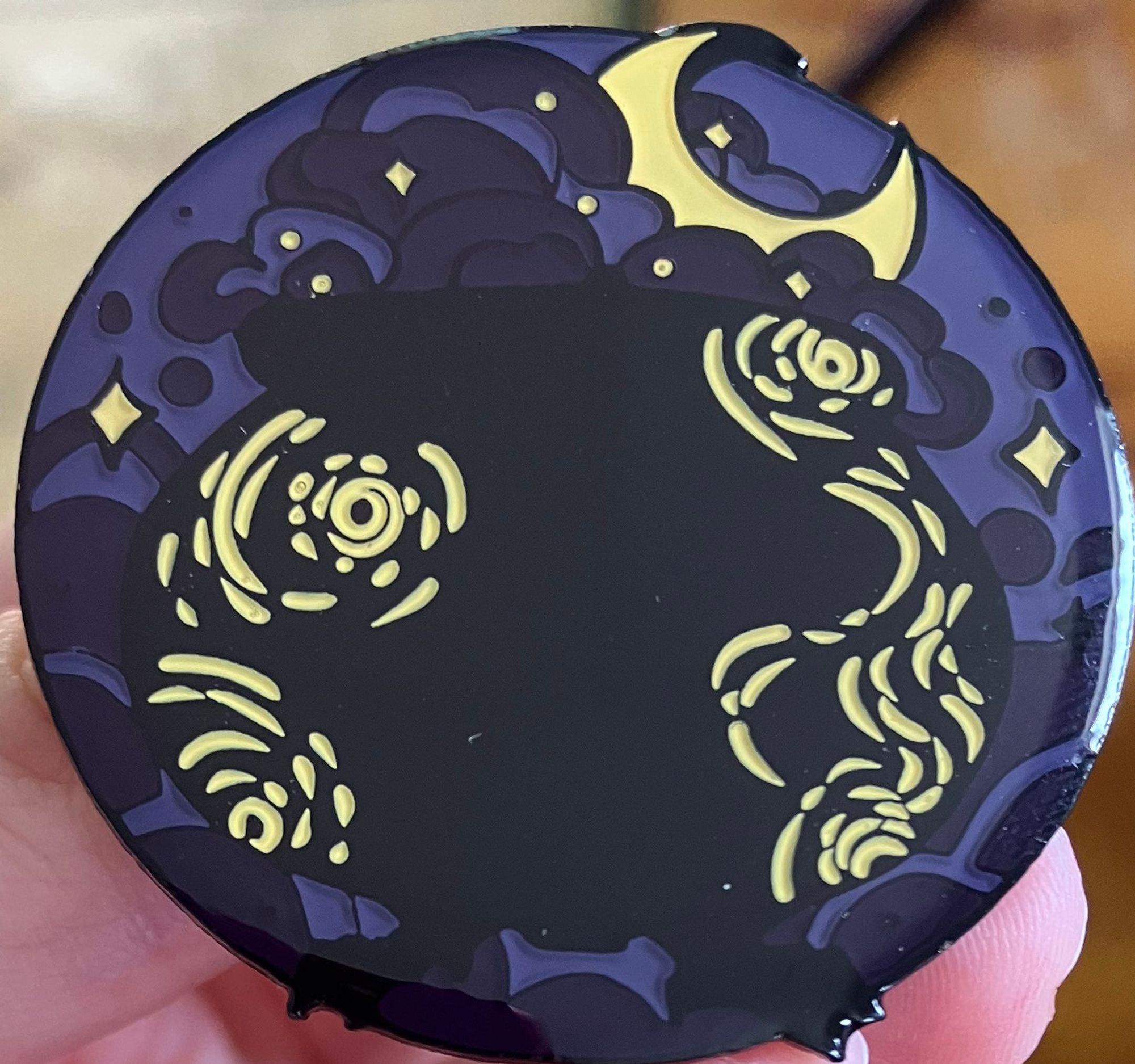 An enamel pin of a cauldron overflowing with a moon in the brew. It is as if the night is flowing out of it.