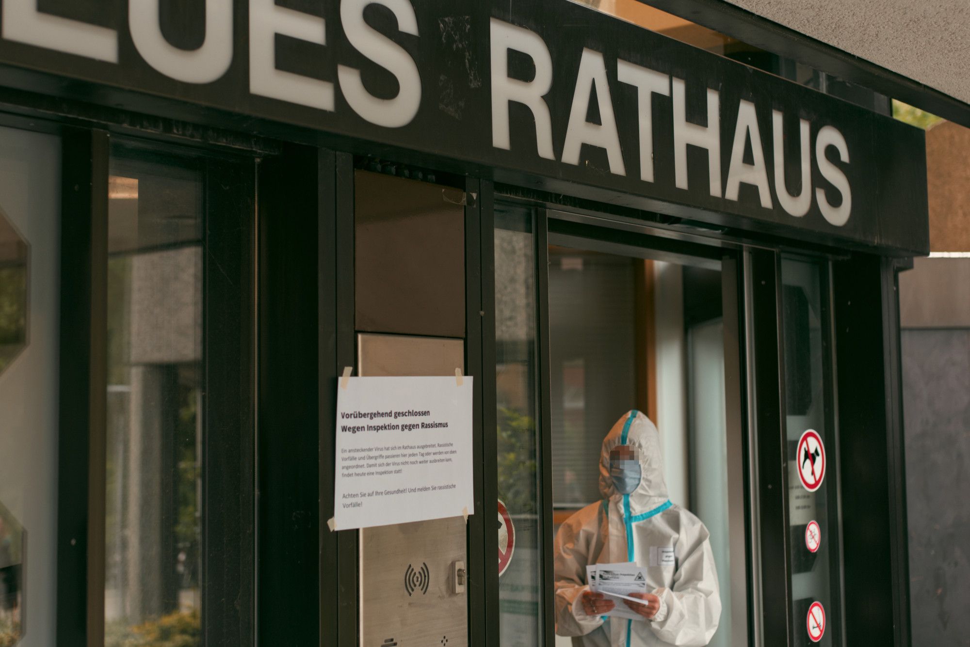 Eine Person im weißen Schutzanzug mit Flyern in der Hand in der Eingangstür zum Neuen Rathaus. Links daneben klebt ein Zettel am Türrahmen, der die vorrübergehende Schließung des Rathauses wegen einer Inspektion gegen Rassismus verkündet.