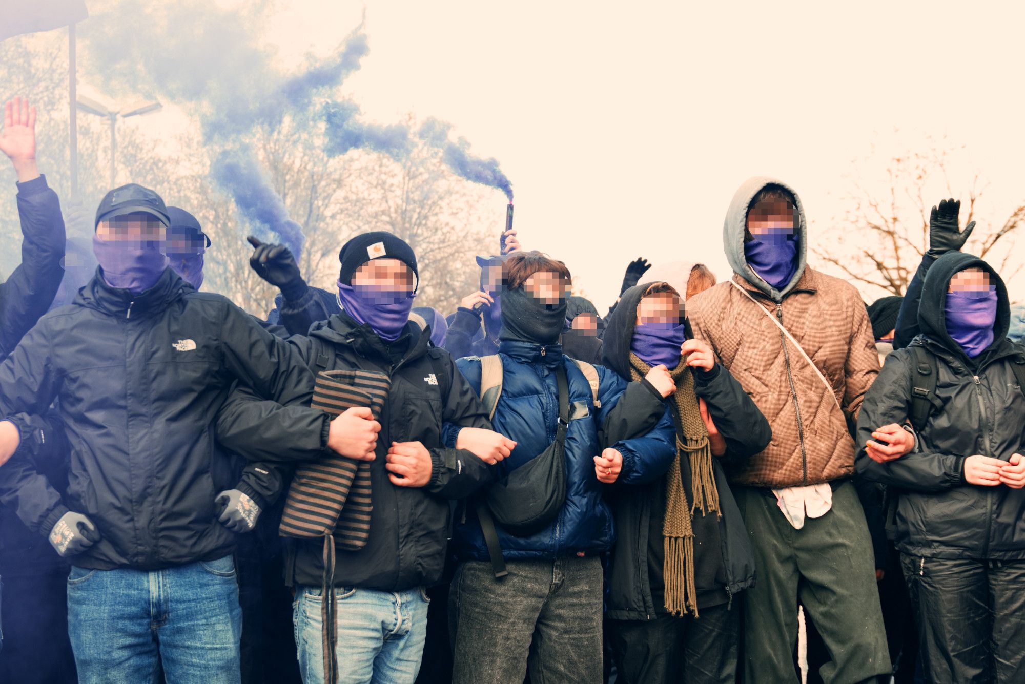 Eine Gruppe vermummter Menschen, die untergehakt eine Blockade bilden. Im Hintergrund wird lilafarbener Rauch gezündet.