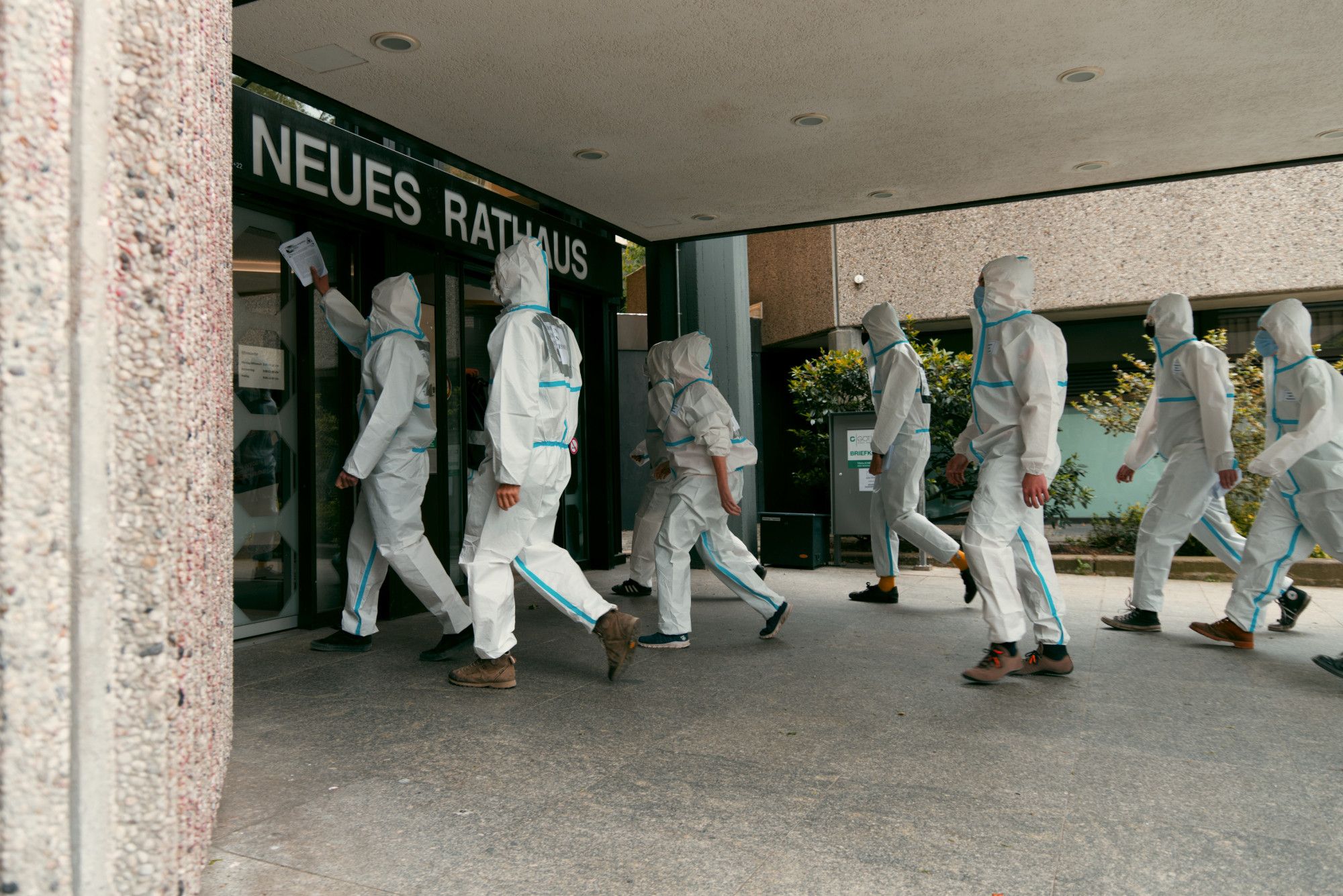 Eine Gruppe von in weißen Schutzanzügen gekleideten Aktivist*innen geht entschlossen auf die Eingangstüren des Neuen Rathaus zu. Eine Person links hält einen Zettel hoch