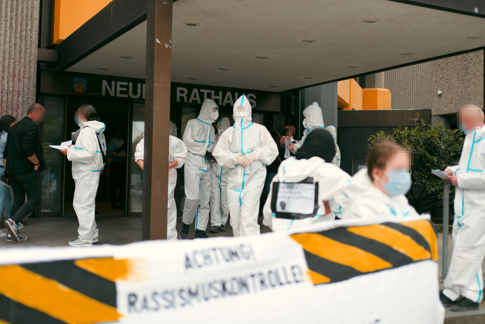 Diverse in weißen Schutzanzüge gekleidete Personen stehen vor dem Eingang des Neuen Rathaus und verteilen u.a. Flyer an unbeteiligte Bürger*innen. Vorne ein weißes "Achtung! Rassismuskontrolle!" Transparent mit einem schwarz-gelben Warnband.