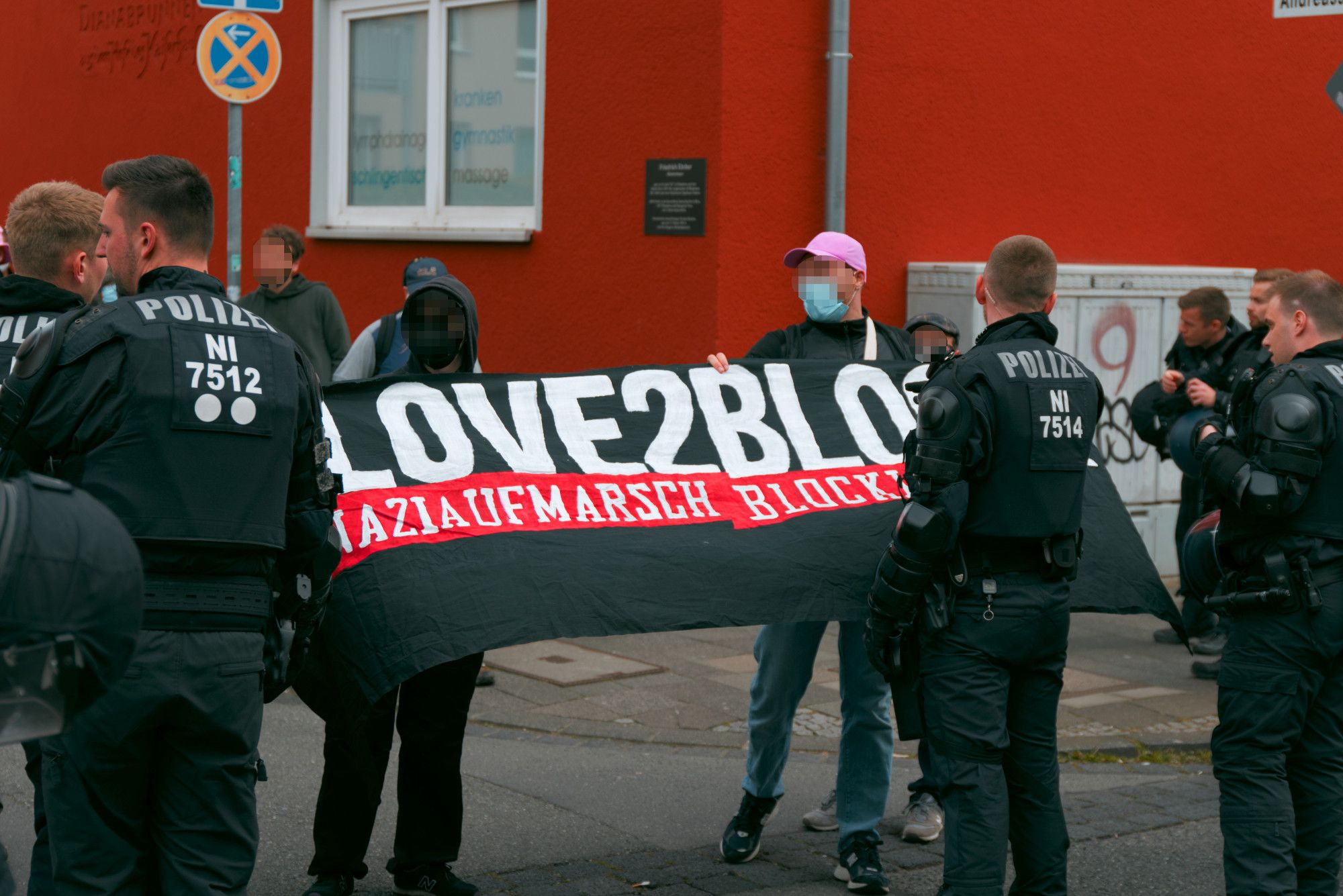 Antifas hinter einer Polizeikette am Rand der Route von Querdenken. Sie halten ein schwarz-rotes "Love2Block. Naziaufmarsch blockieren." Transparent, das teilweise verdeckt ist.