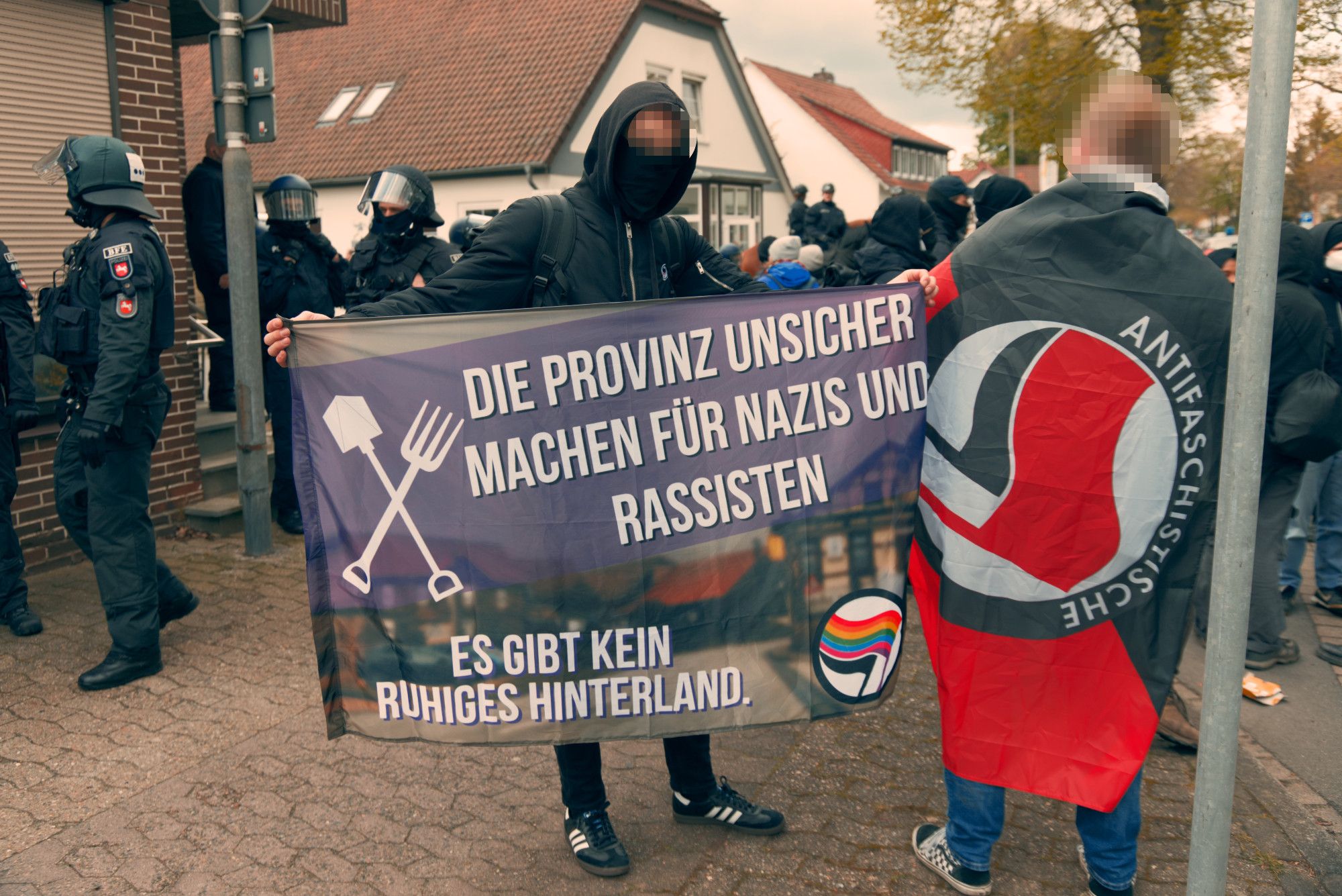 Eine Person mit einer "Die Provinz unsicher machen für Nazis und Rassisten. Es gibt kein ruhiges Hinterland." Fahne neben einer Person mit einer schwarz-roten Antifa-Fahne. Beide stehen vor einer blockierten Kreuzung.