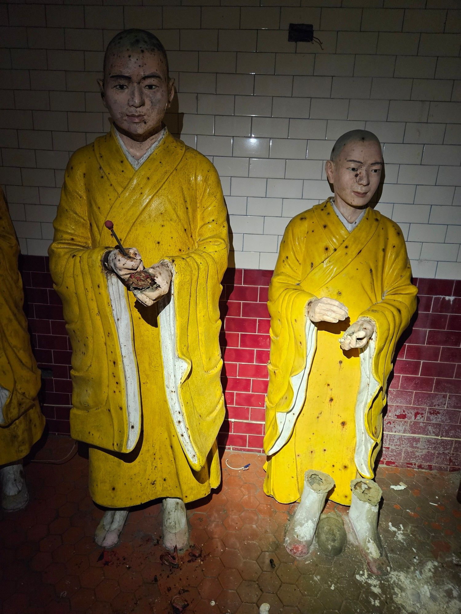 Statues of two monks evoking the "uncanny valley" effect. They have yellow robes and are covered with corrosion. The one on the right has also been separated from his feet.