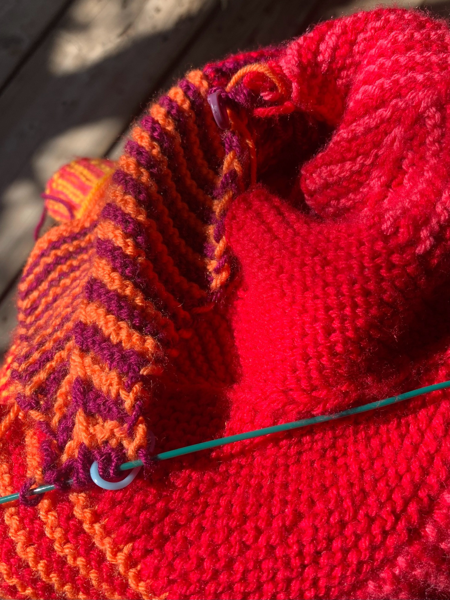 a pile of the knitting on juni’s lap in the sun. the primary colors of the afghan in this shot are red, orange, purple, and pink. there are a few stitches of active purple stitches and a blue stitch marker on a teal knitting cable. 