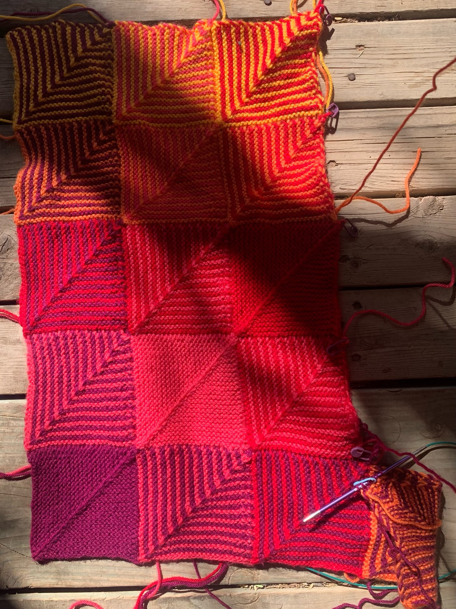 a section of the hue shift afghan in progress in a sunbeam on juni’s porch. the afghan is a series of rows and columns of squares knit on to each other and in this section the colors are purple, pink, red, orange, and yellow. some of the squares are solid colors but most squares are striped using two of the colors mentioned in each square. 