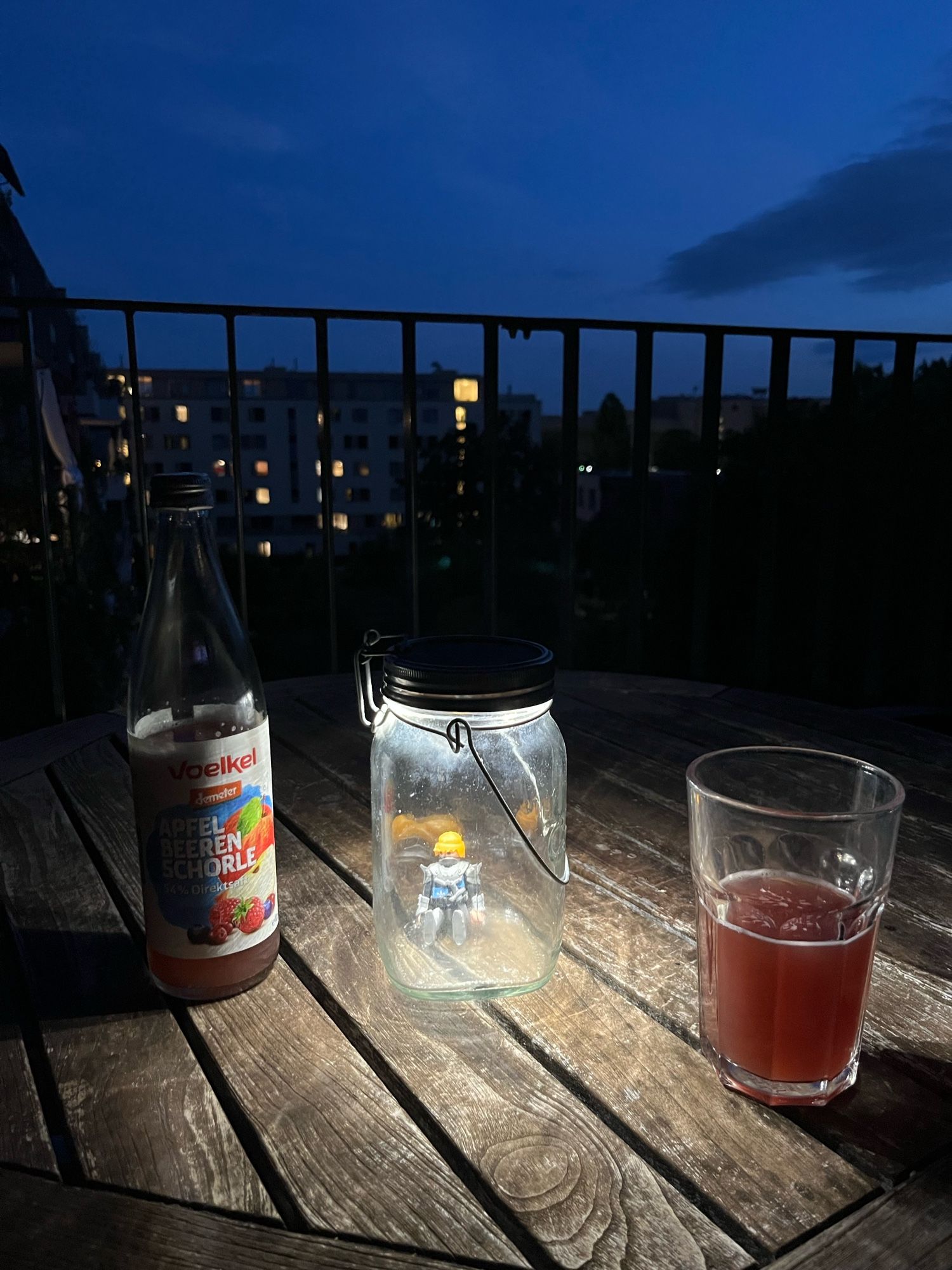 Ein Tisch mit einer Flasche und einem Glas Apfelbeerenschorle sowie einem Solarlicht auf einem Balkon, dahinter der Umriss des Tempelhofer Flughafengebäudes