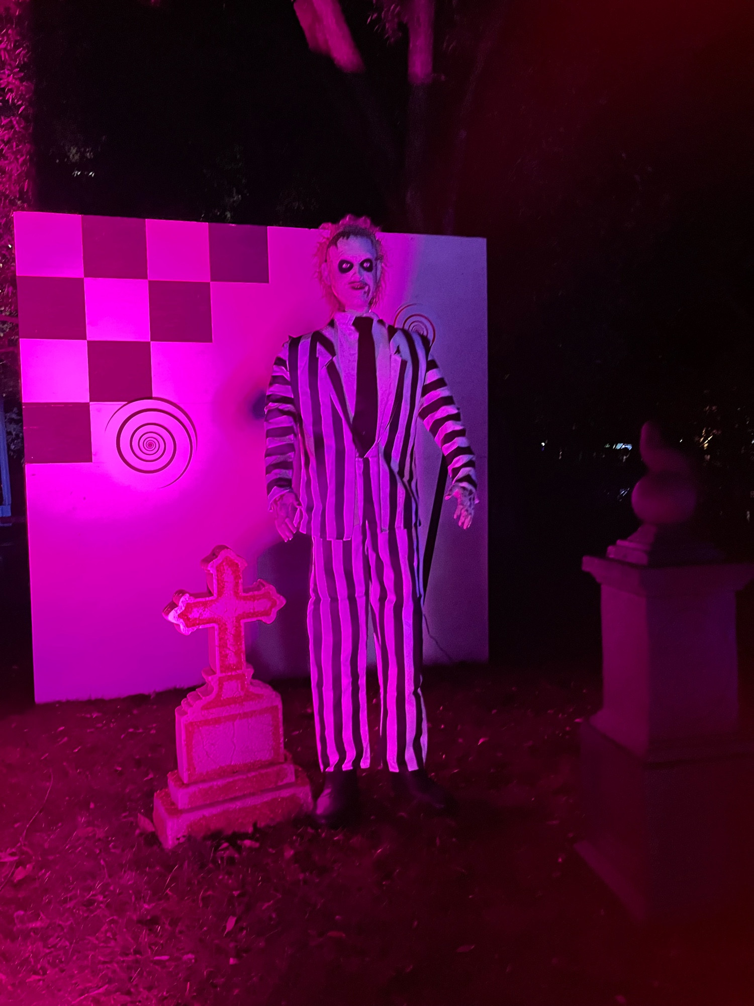 Beetlejuice is standing next to a grave in front of a cardboard background in a forest trail.