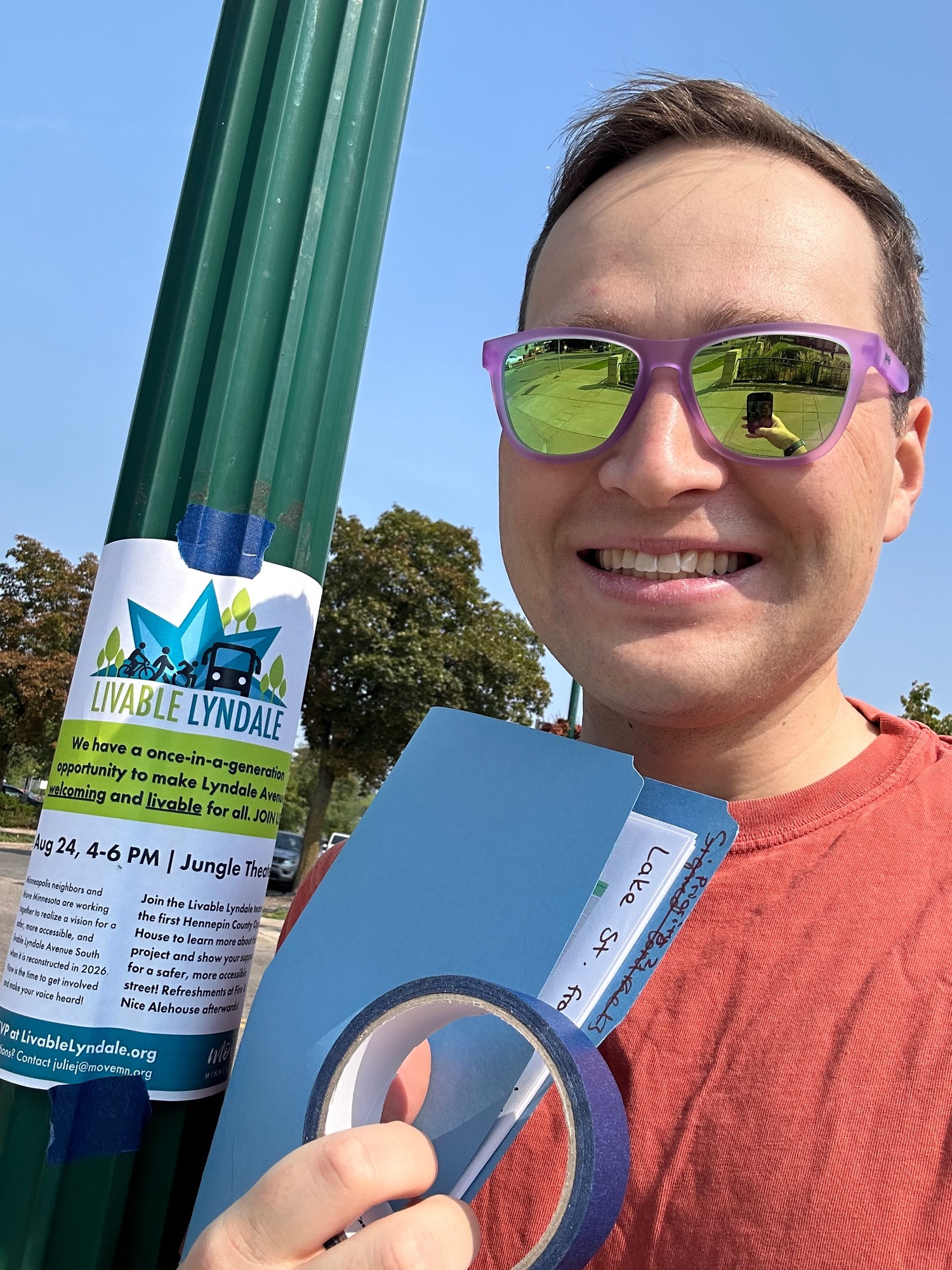 Brian smiling with sunglasses taking a selfie next to a streetlight pole with a flyer taped onto it while holding a folder with more fliers and a roll of painter tape.