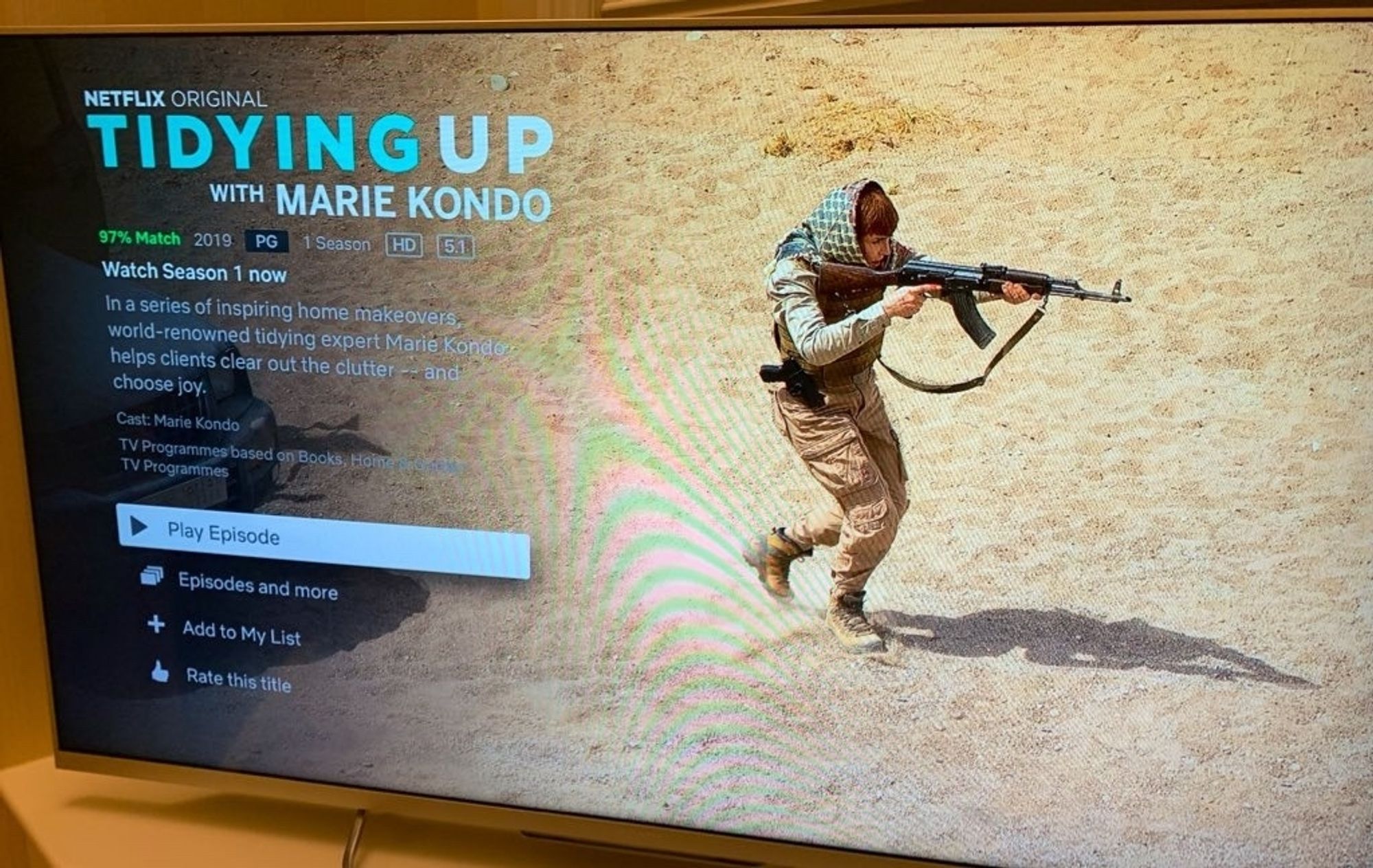 A picture of a television displaying a glitched out Netflix so that it says that the show is tidying up with Marie kondo but the picture is a preview from something else, not sure what but it's a person stalking the desert with an AK-47