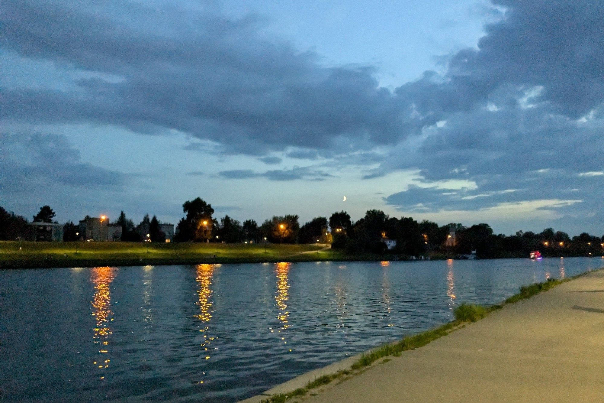 crescent moon against a pale blue sky spanned by dark blue clouds, trees along the far riverbanks dotted with street lamps glowing orange, reflecting in the ripples