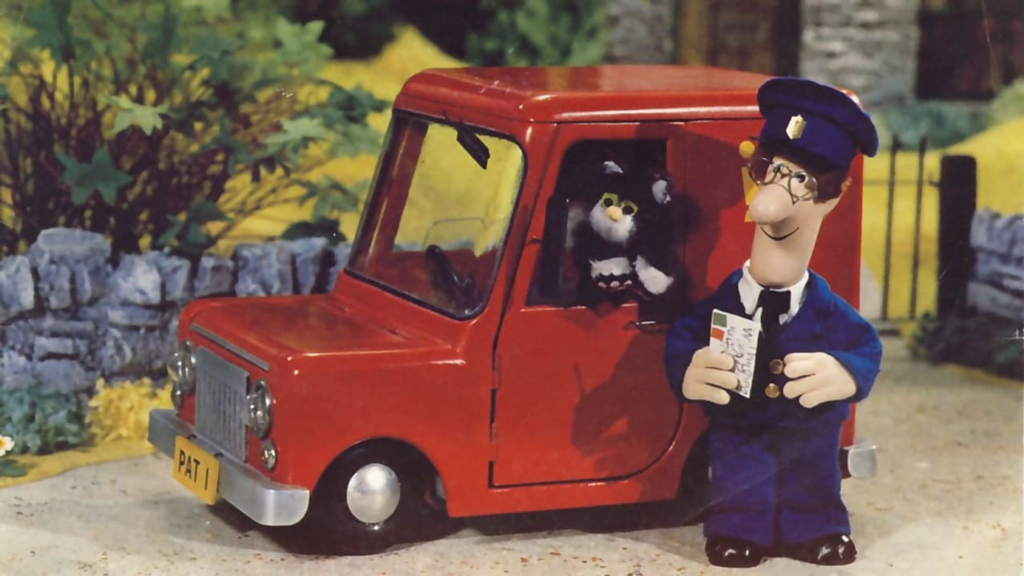 A still from the stop motion animated Postman Pat. The eponymous Pat stands in front of a red Royal Mail van with a tall windscreen and cabin, his cat leaning out the window.