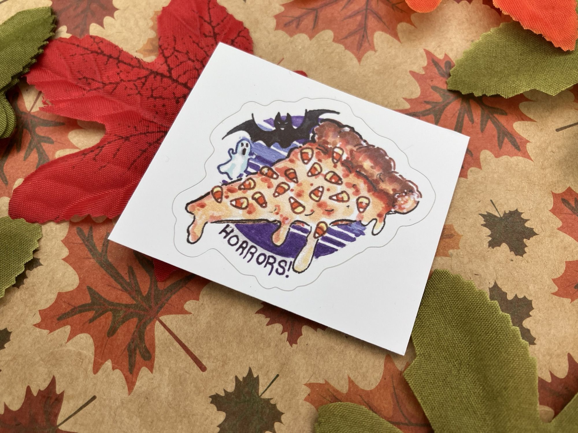 a sticker of my drawing of a slice of pizza covered in candy corn captioned "horrors!" with a tiny bat and ghost looking at it as it goops all over. it's on a background with fall leaves printed on paper and some silk faux leaves scattered around.