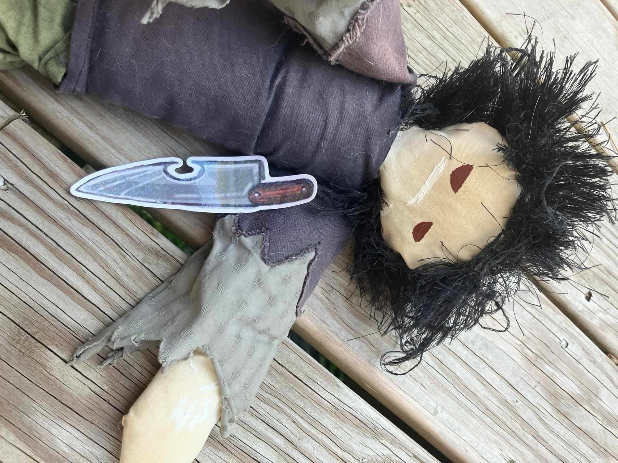 a ragdoll plush of my protag Grey (i made it) lying on wood planks looking sullen, with the sticker knife against his chest.