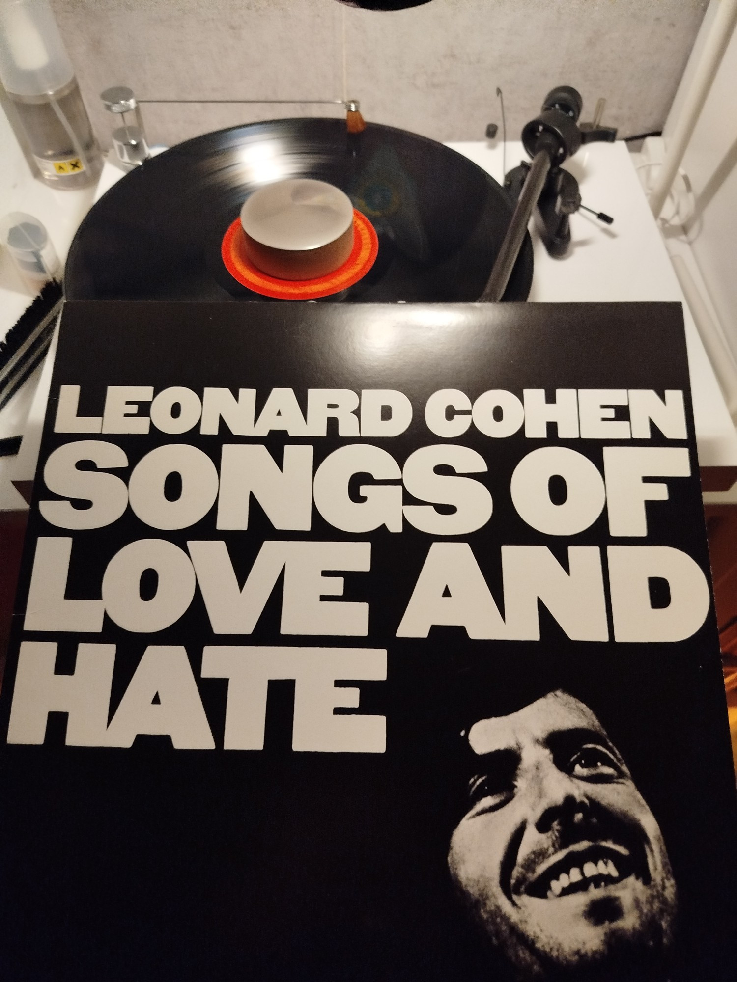 Das Bild zeigt ein Plattencover von Leonard Cohens Album "Songs of Love and Hate". Das Cover ist schwarz-weiß gehalten und zeigt den Titel des Albums in großen, weißen Buchstaben, die den oberen Teil des Covers dominieren. Unten rechts befindet sich ein Porträt von Leonard Cohen, der lächelnd nach oben schaut.

Im Hintergrund sieht man einen Plattenspieler, auf dem eine Schallplatte aufgelegt ist. Die Schallplatte hat ein auffälliges, orangenes Label in der Mitte. Der Tonarm ist bereits über der Schallplatte positioniert, was darauf hinweist, dass die Platte entweder gerade spielt oder bereit ist, abgespielt zu werden.

