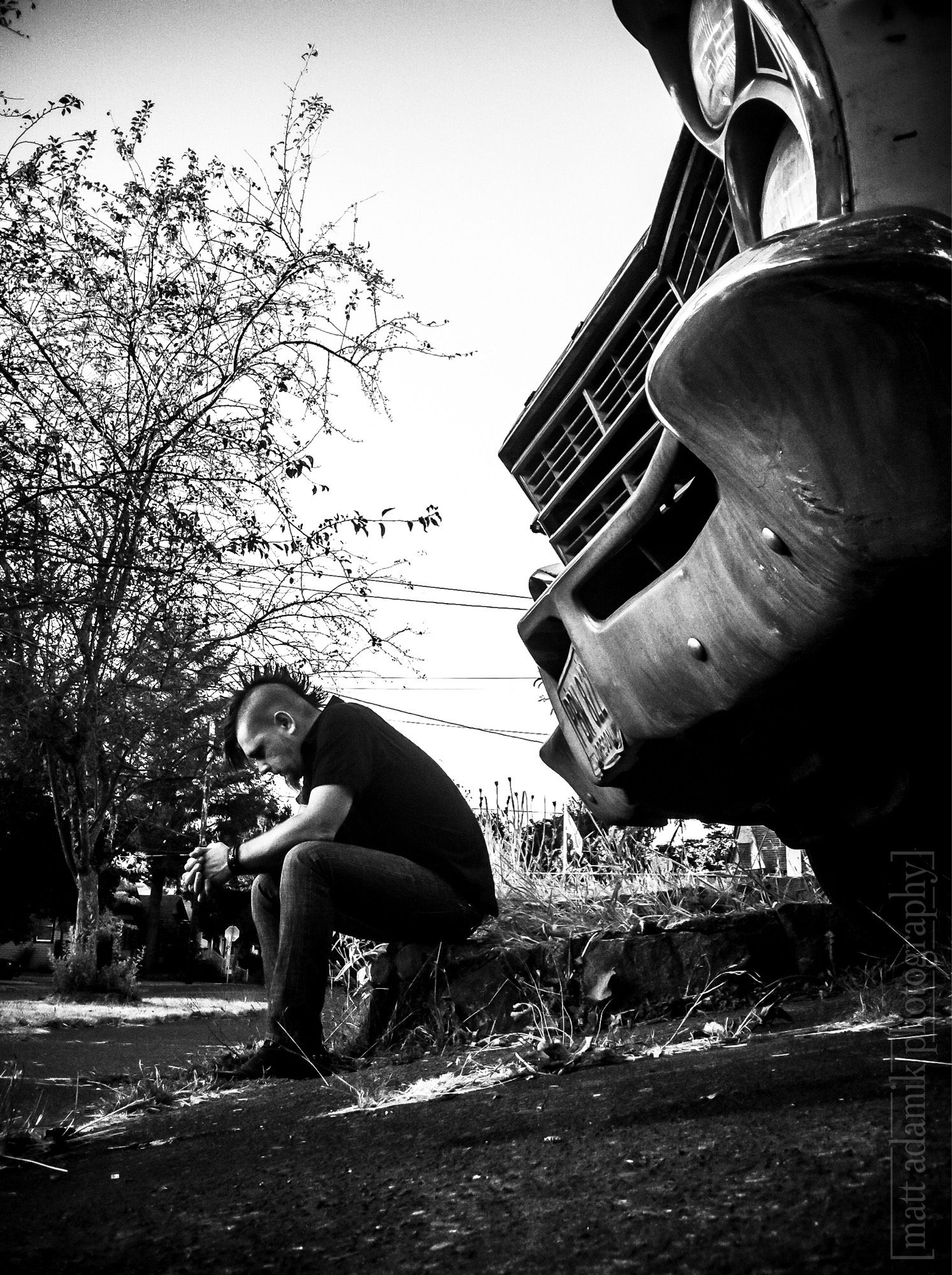 Self portrait black and white photograph of the photographer sitting on a retaining wall at the end of a driveway with the bumper of a 60’s Ford Galaxy framing the foreground.