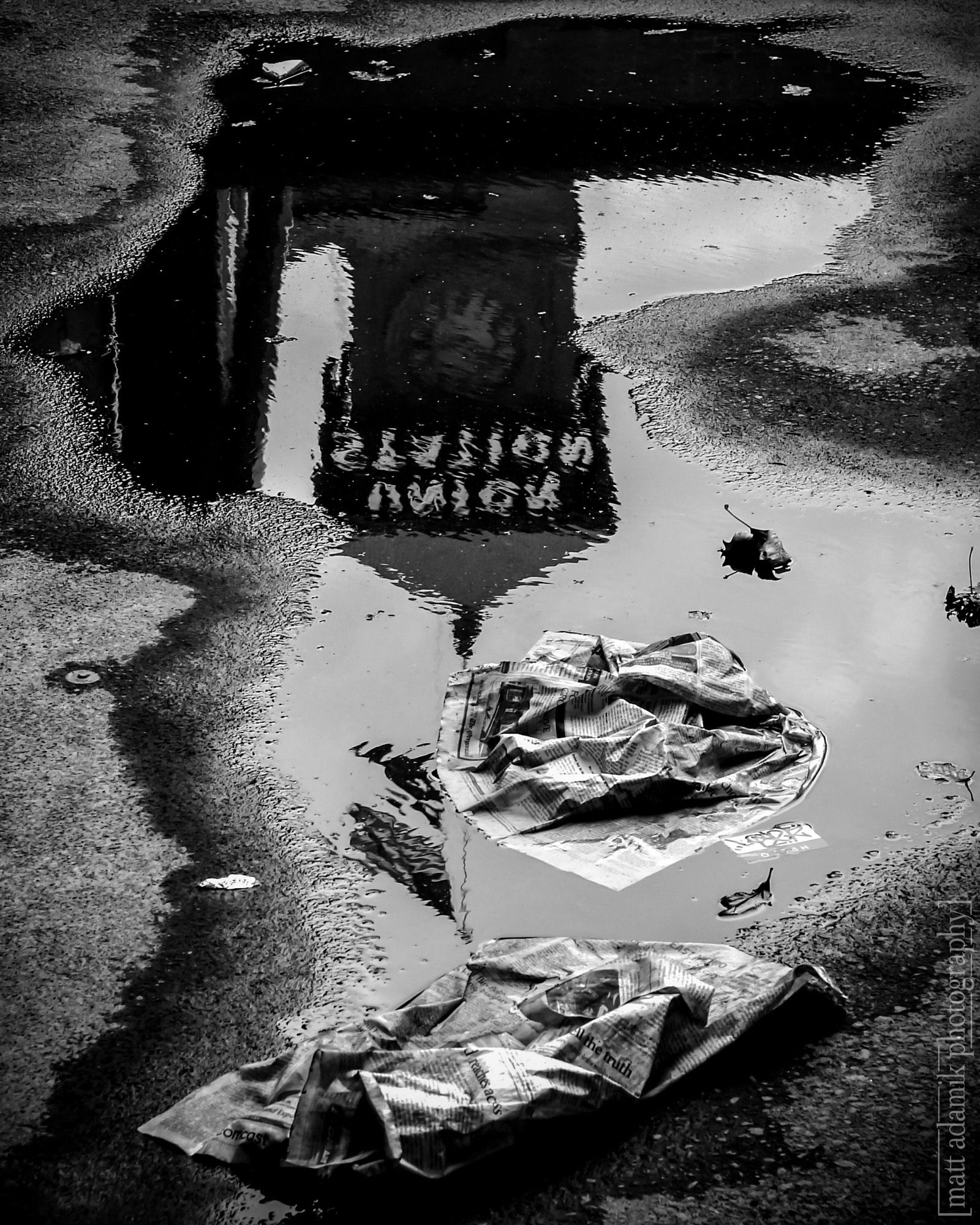 Black and white photograph of the upside down reflection of the tower at Portland’s Union Station train station in a puddle with some crumpled wet newspapers in the foreground.