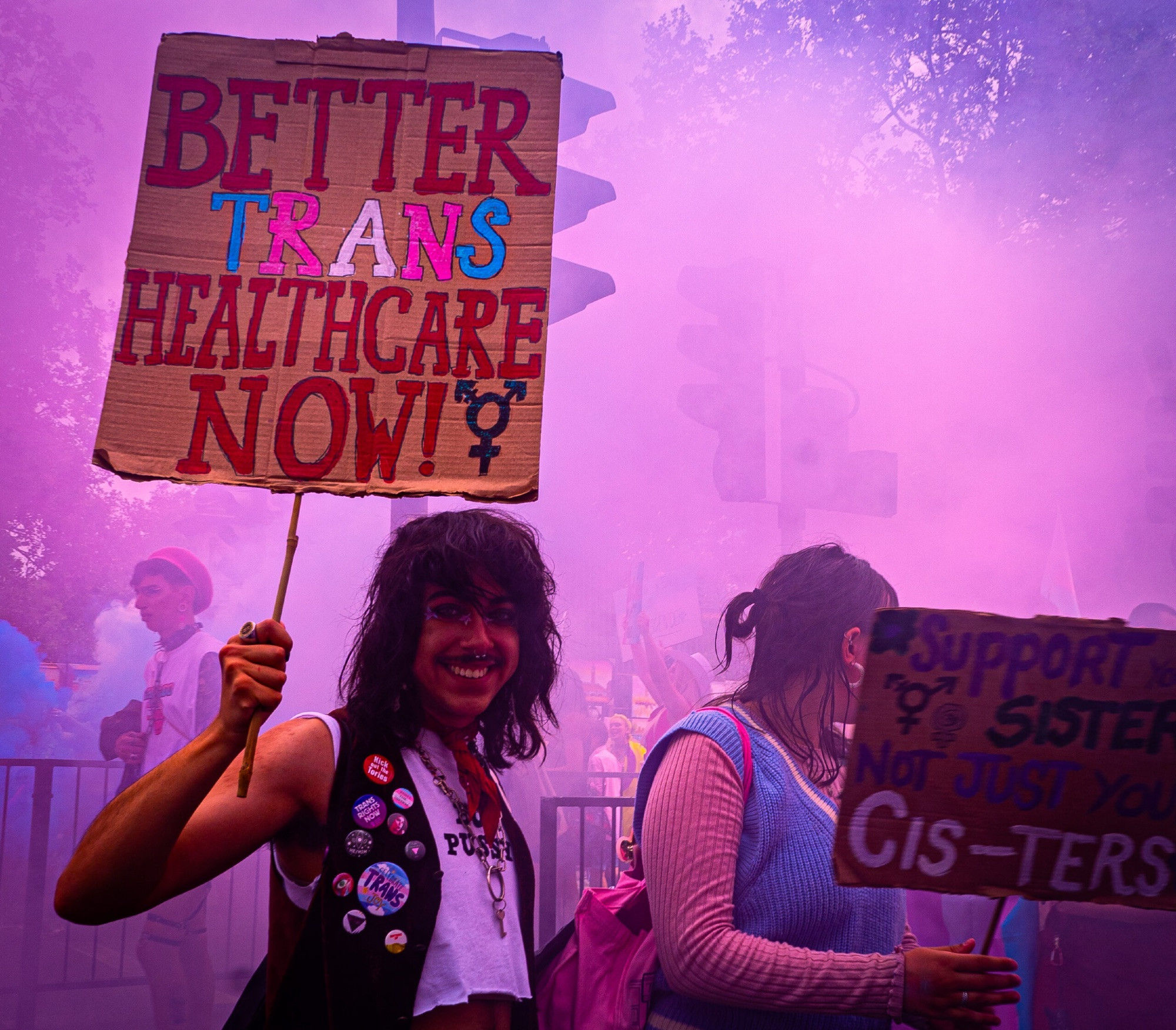 pic from the article of protesters in the UK sign reads "better trans healthcare now" and "support your sisters not just your cis-ters"