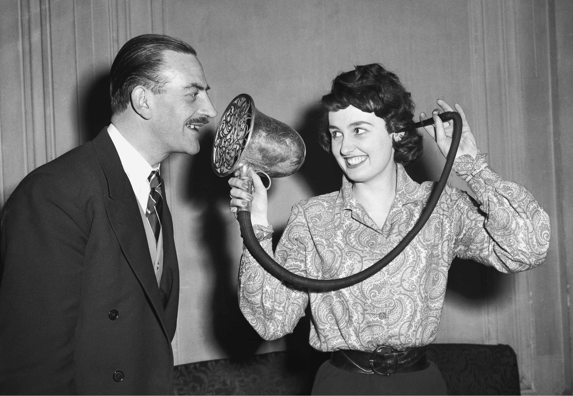 mid twentieth century black and white picture of two people, both dark haired white people, one man and one woman. they are smiling. the woman has a tube jammed in her ear. it is attached to a hose attached to what looks like a giant flashlight or hair dryer or something. it’s an over complicated ear horn. if your grandmother bought this, she was still mad about it in 1991.