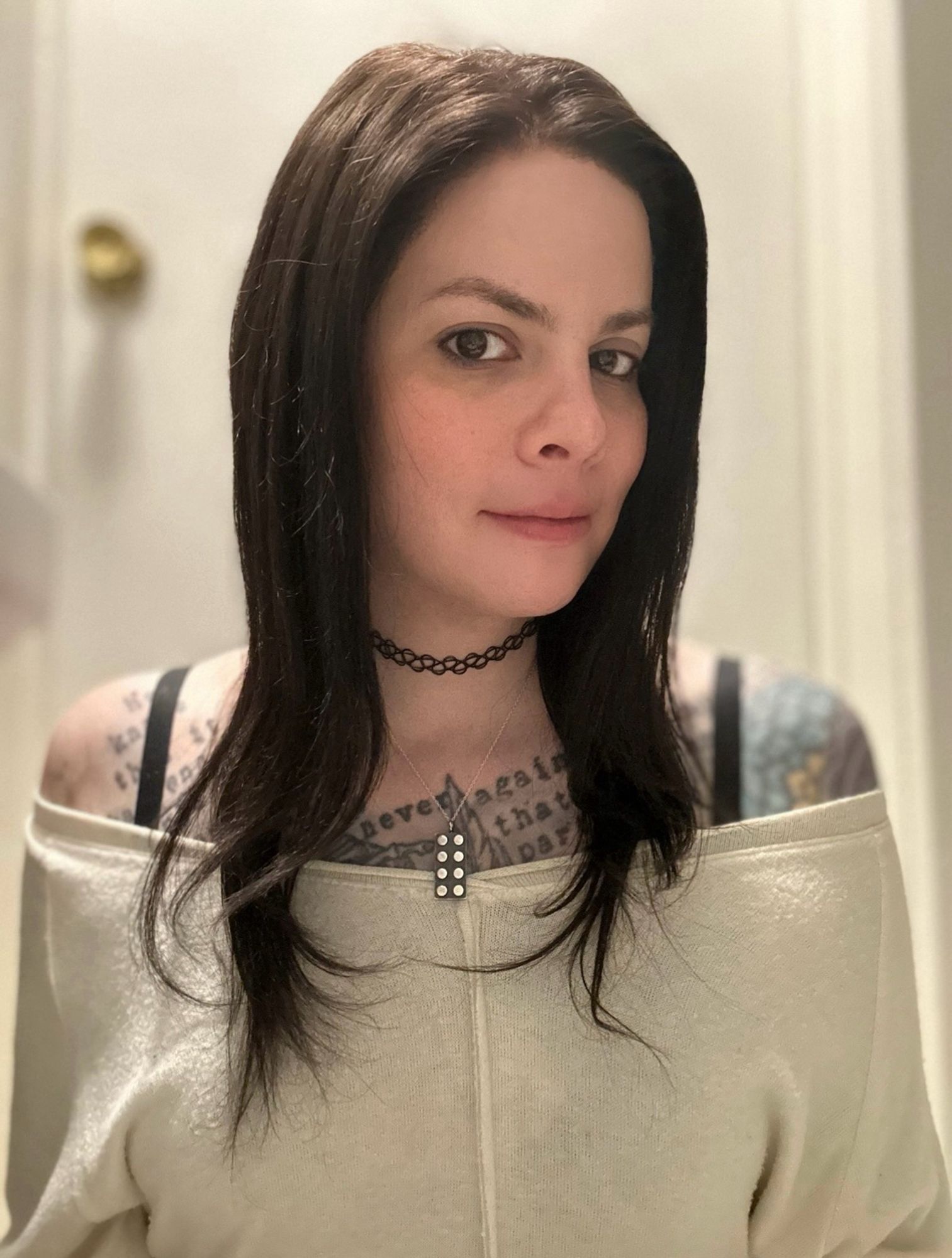 brunette white woman with flatironed hair. white off the shoulder top. black bra straps visible. one of her shoulders is tattooed blue. the other is now fairly bare. her chest has a lot of black typewriter text. steel 2x4 lego brick necklace. she is smirking to let you know she wouldn’t break her vegetarianism to eat a transphobe. uwu.