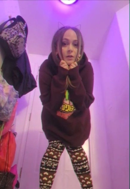 Low angle of a girl dressed in oversized brown hooded sweatshirt with decorative leggings with hands holding her hair over her cheeks, with a neutral expression on her face. An overhead ambient light shines above her head.