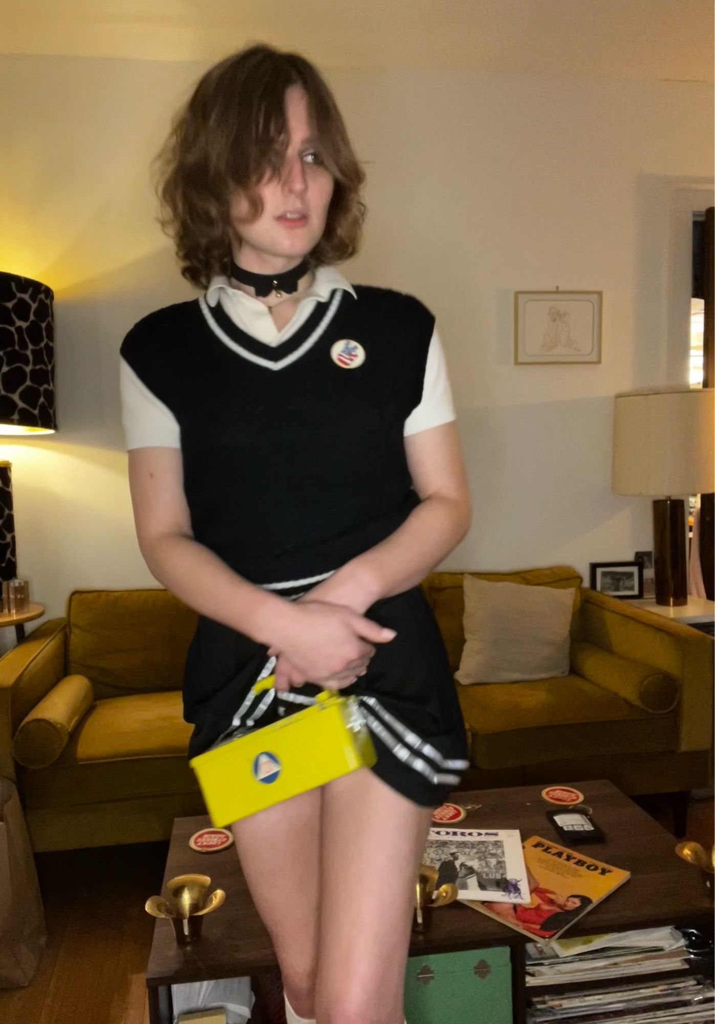 harper wearing white knee highs holding the front of her skirt with a yellow civil defense Geiger counter over her bulge she's wearing a black sweater vest with white stripes and a white chevron accenting the v neck and a white polo. She is also wearing a black collar and a peace sign hand american flag pin in the background her messy coffee  table and her couch