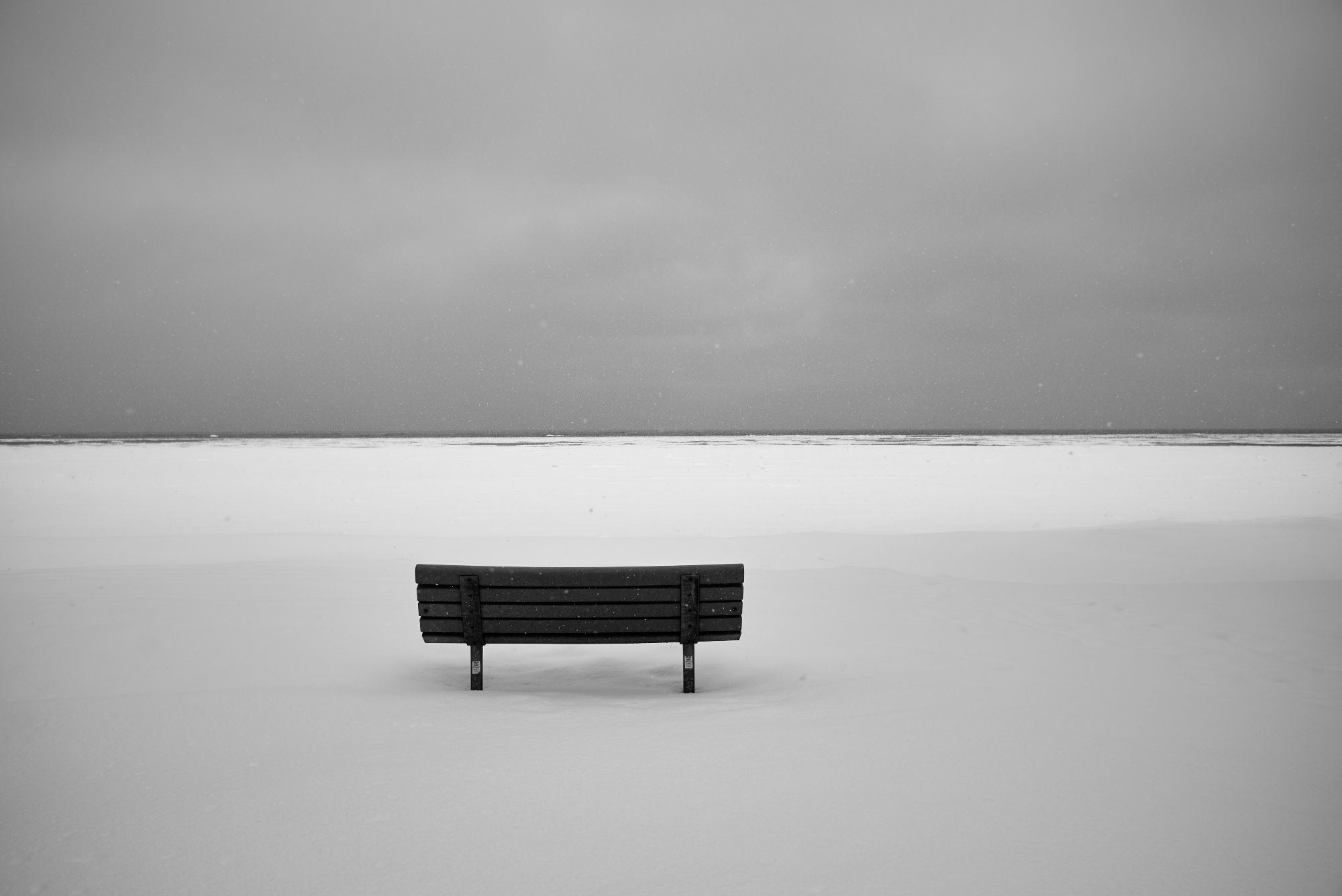 A black and white photo of bench in the snow with snow almost to the horizon.