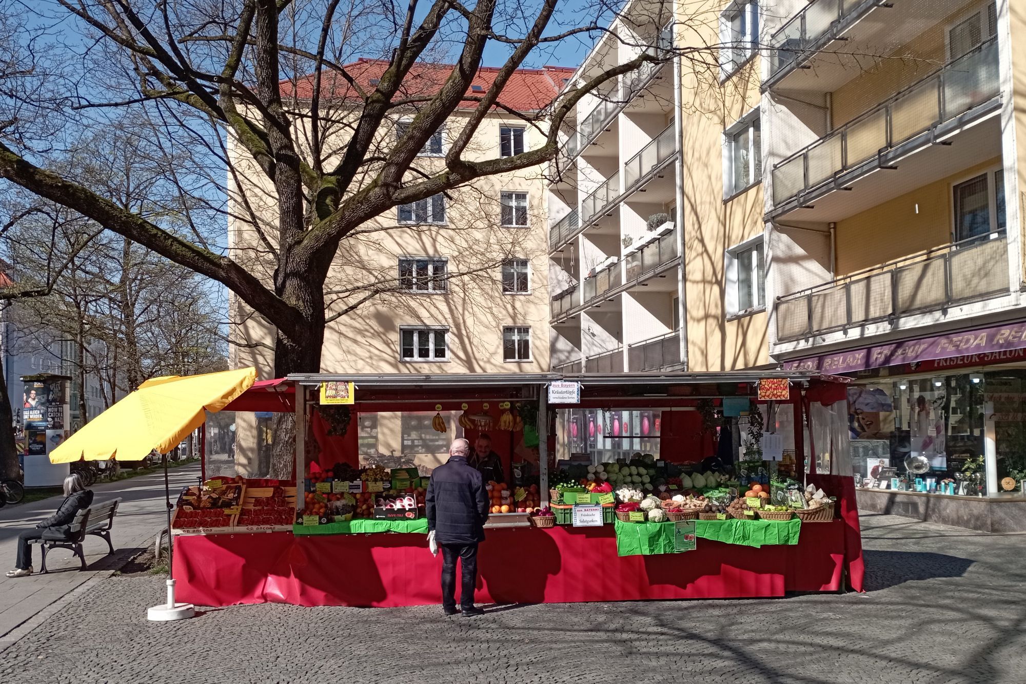 Obst- und Gemüsestand am westlichen Rand des gepflasterten Hohenzollernplatzes bei klischeehaftem Frühlingswetter. Im Hintergrund vierstöckige Wohnhäuser