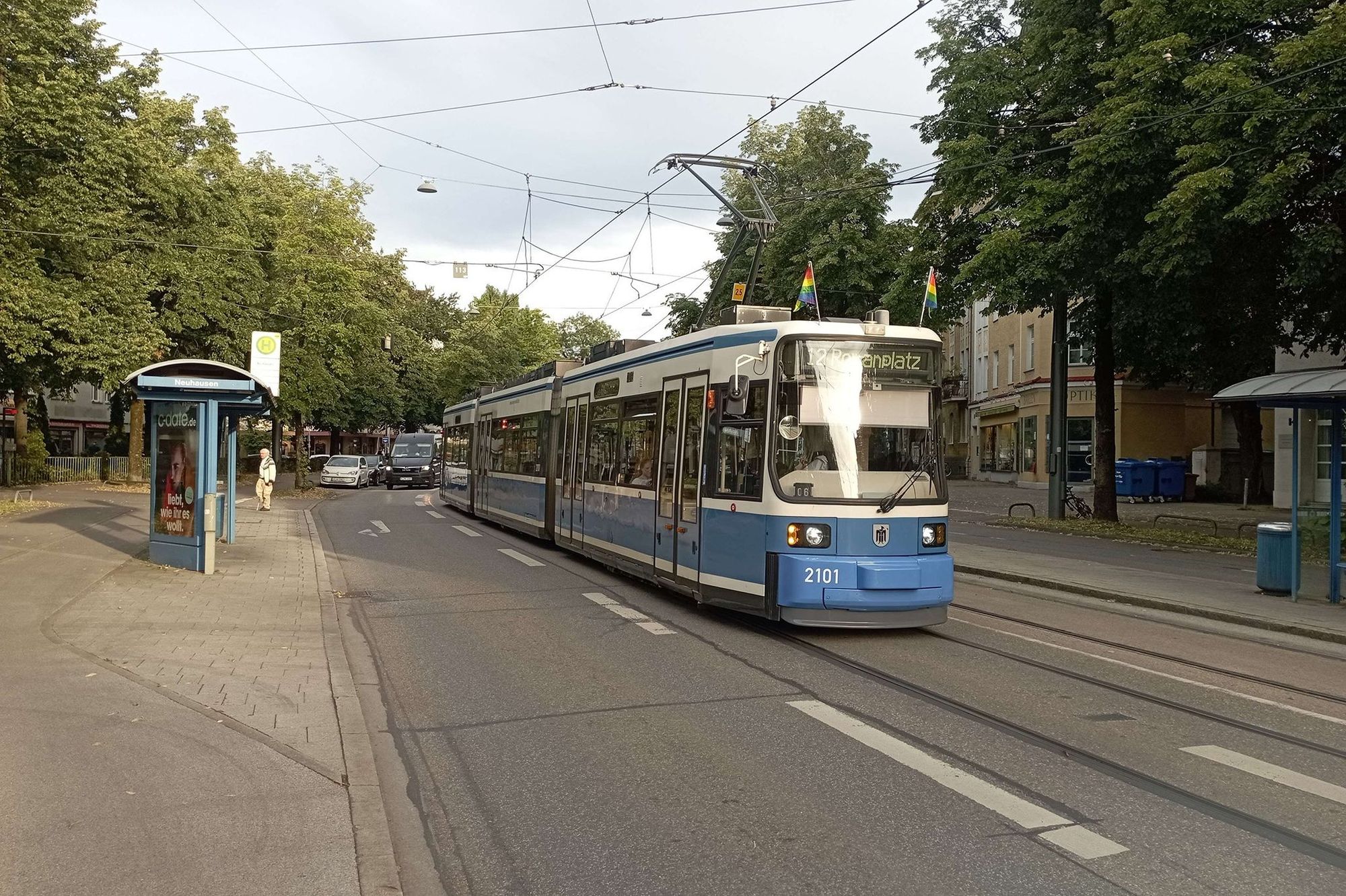 [Querformat/Farbe] Weißblaue, nicht redesignte Tram der MVG vom Typ GT6N/R 1.1 mit Priede Month-Beflaggung auf dem Dach an der H Neuhausen Richtung Romanplatz. Die Frontschürze trägt das Blau, das die redesignten R 1.1 haben.
Links Haltestellenhäusl, dazwischen Straße, dir für Autos Rot für Fahrgastwechsel hat.