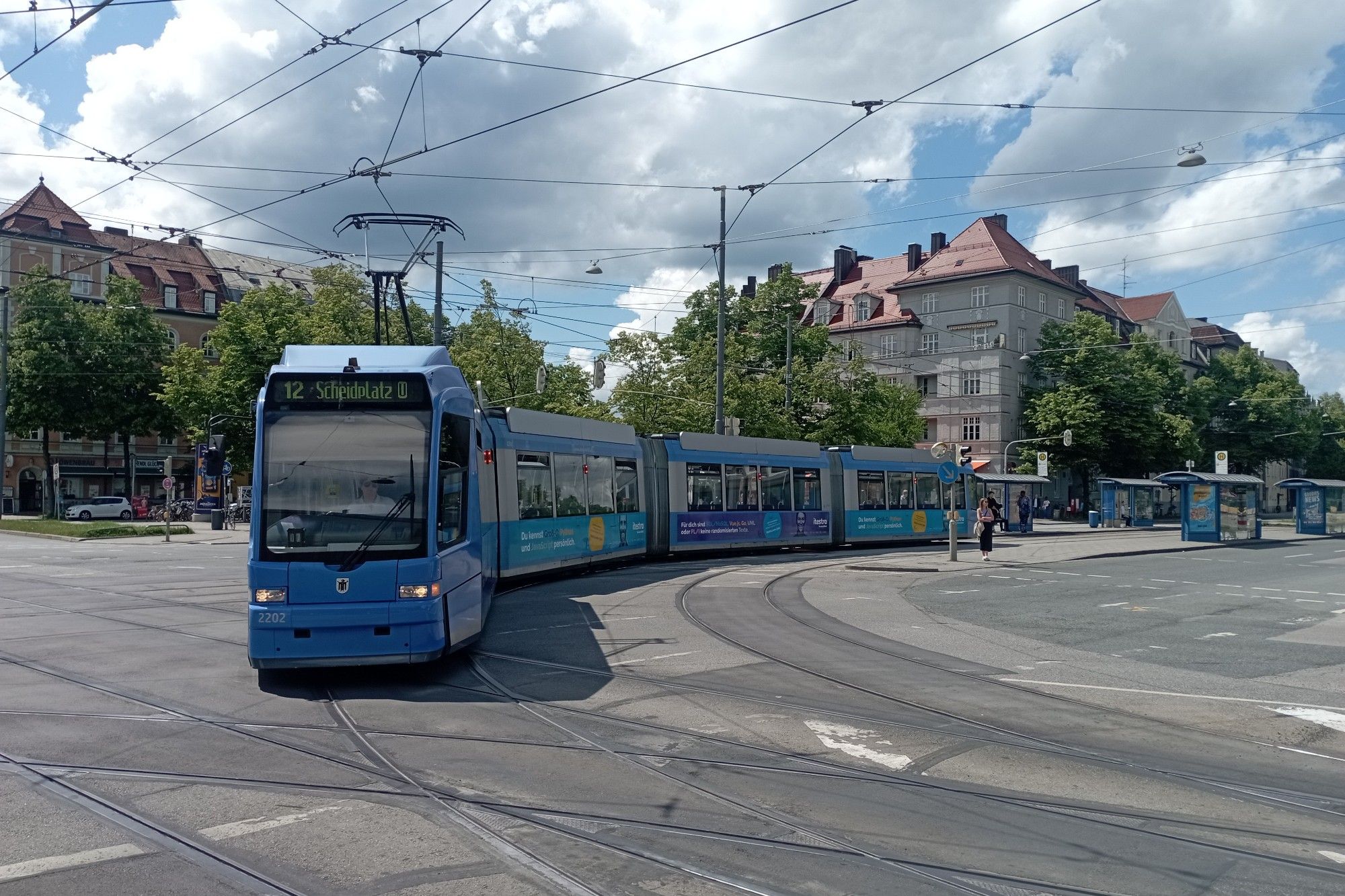 Vierteilige Tram vom Typ R3 in Blau-Silber biegt bei strahlendem Sonnenschein am Leonrodplatz von der Dachauer in die Schwere-Reiter-Straße ab.