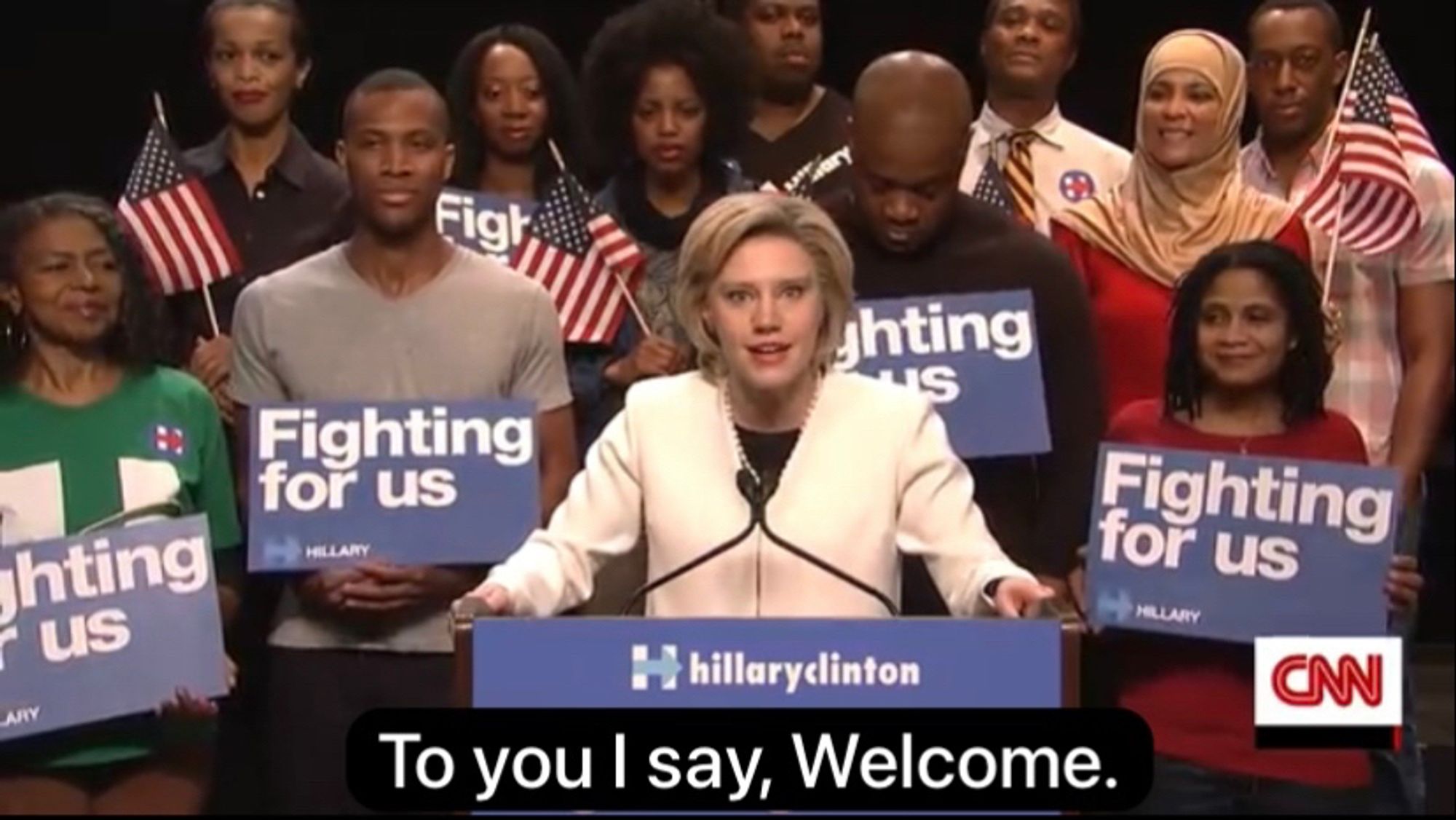 Image of Kate McKinnon as Hillary Clinton on Saturday Night Live in 2016 standing at a podium. Behind her are actors playing supporters who hold up American flags and signs that say "Fighting for us". The podium says "hillaryclinton". The subtitle at the bottom of the image says "To you I say, Welcome."