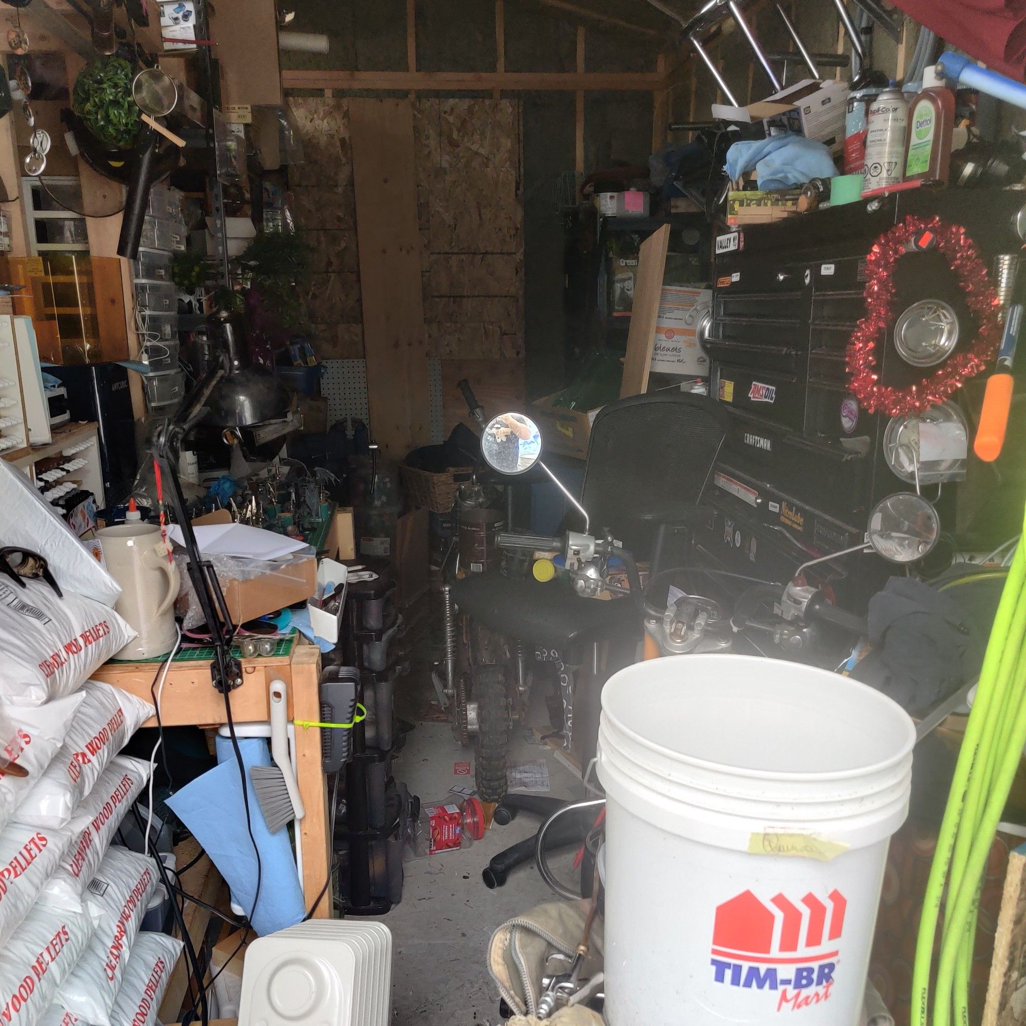 A chaotic view from the doorway to my shed. Visible in this view is a white bucket with the Timber Mart logo. The corner of the paint desk. The mirrors of my Honda CT90/110 motorcycle. A green air hose. An assorted mess of items, which are many and various and not particularly distinguishable.