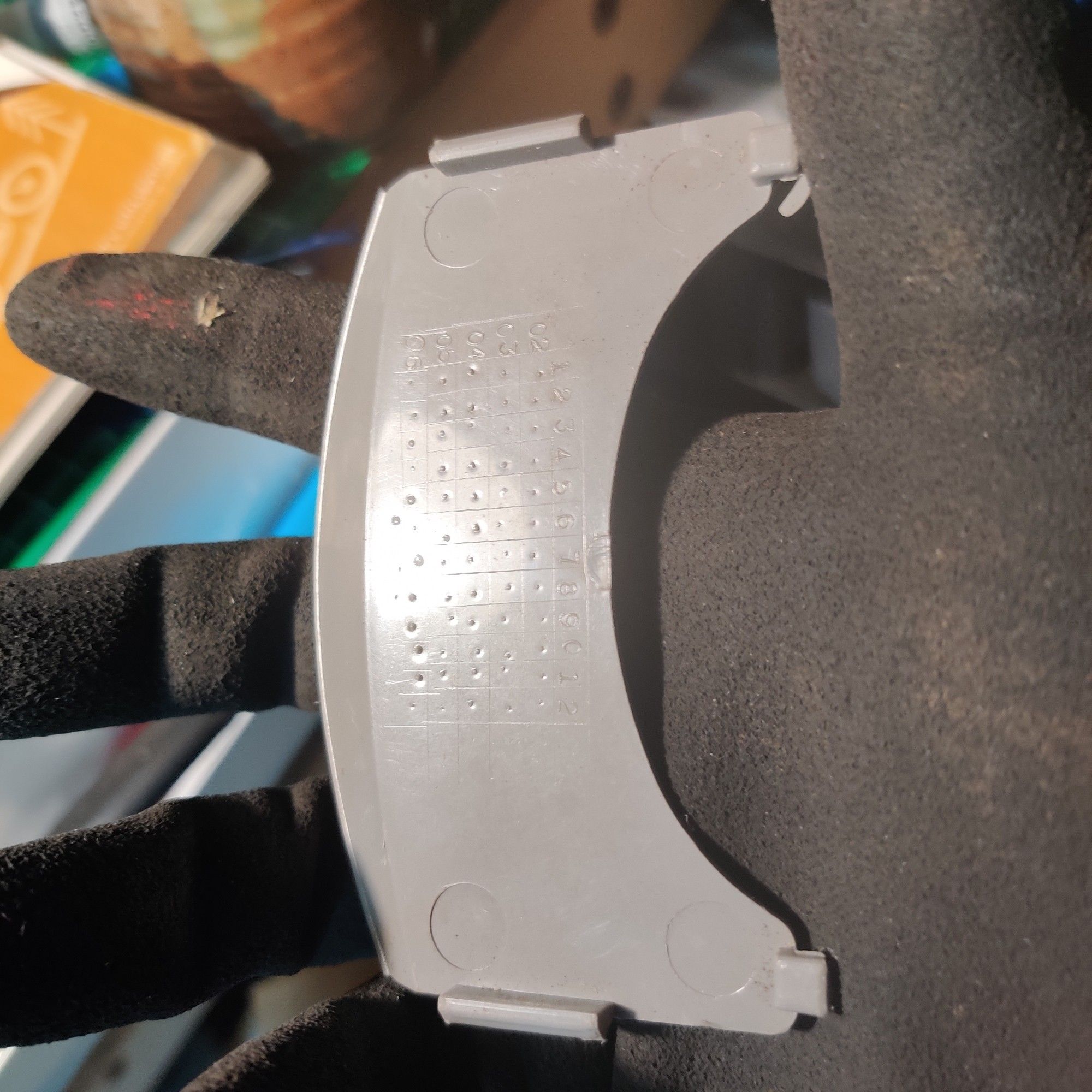 This grey piece of plastic, on my black-gloved hand has been missing for months. It is a cover plate for a Honda CHF50 scooter that fits behind the headlight assembly and protects the light connector from the weather. It is grey and is somewhat crescent shaped with four tabs that fit into slots on the front fairing and leg guard.