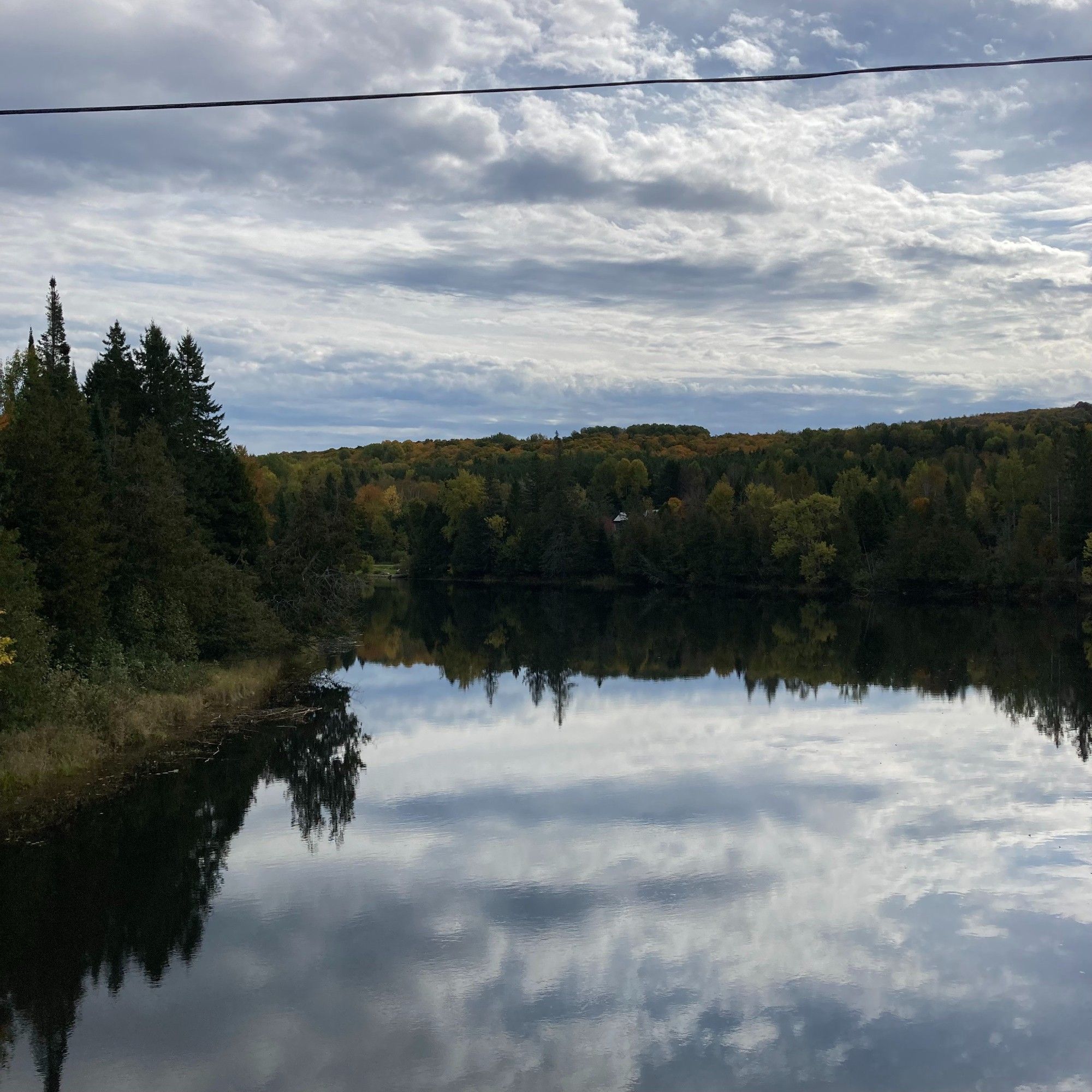 The view from a bridge overlooking water. The sky and clouds are reflected on the water. The trees are changing colour. It is a beautiful fall day.