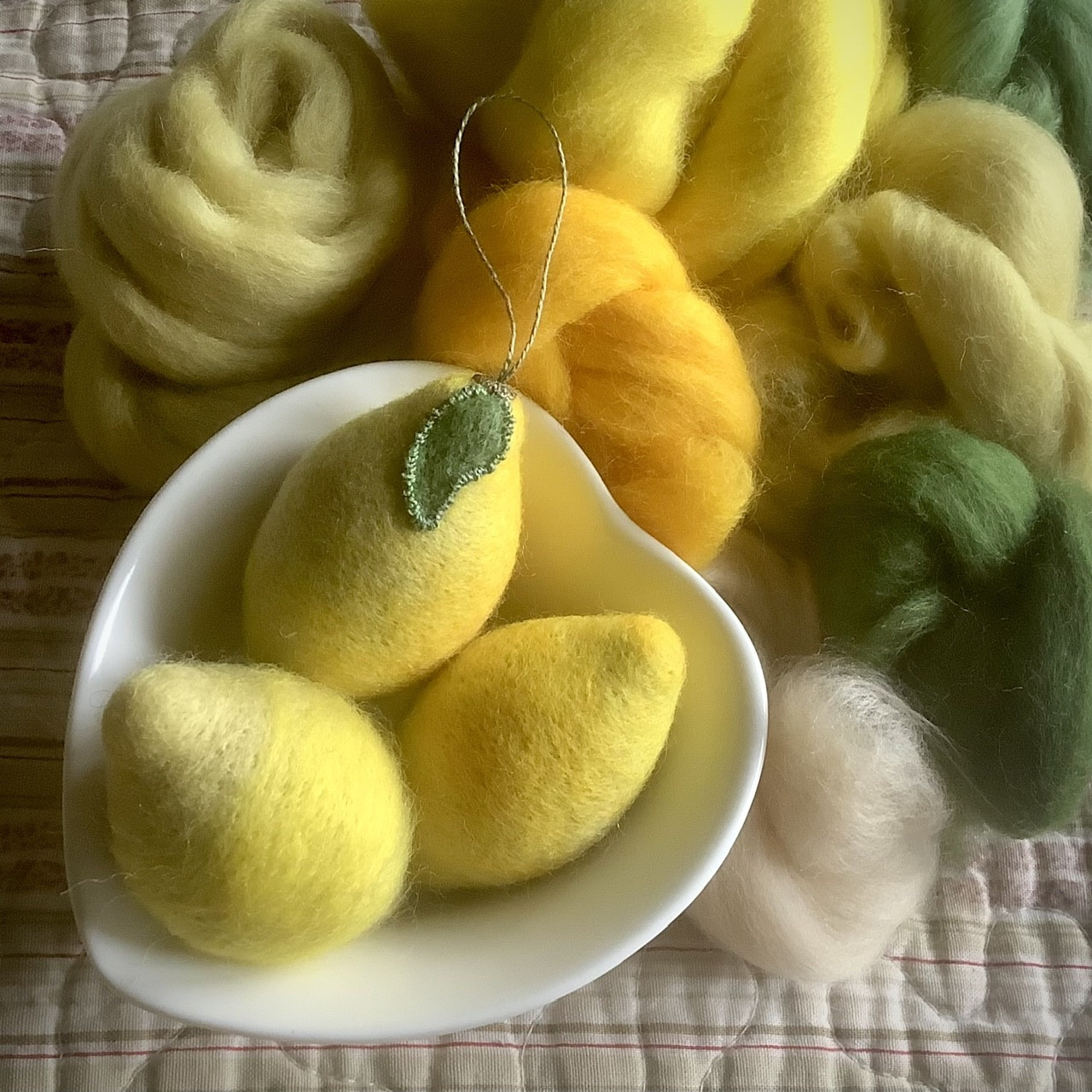 A small white heart shaped bowl with three woollen lemons nestling inside, surrounded by bundles of wool in various yellows and greens, all laid out on a striped quilt top.