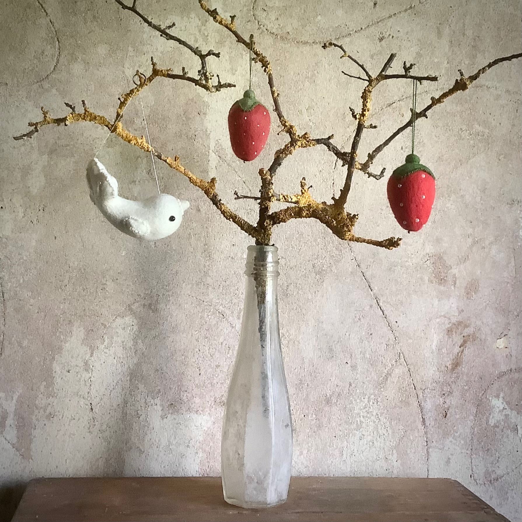 An old tall glass bottle standing on a wooden shelf against a rough plaster interior wall. There is a natural branch station in it with several twiggy offshoots, covered in yellow lichen. A decorative white woollen folk bird hangs from one branch and two red woollen strawberries studded with off white beads hang from others. The atmosphere is plain and sparse so that the decorations stand out.