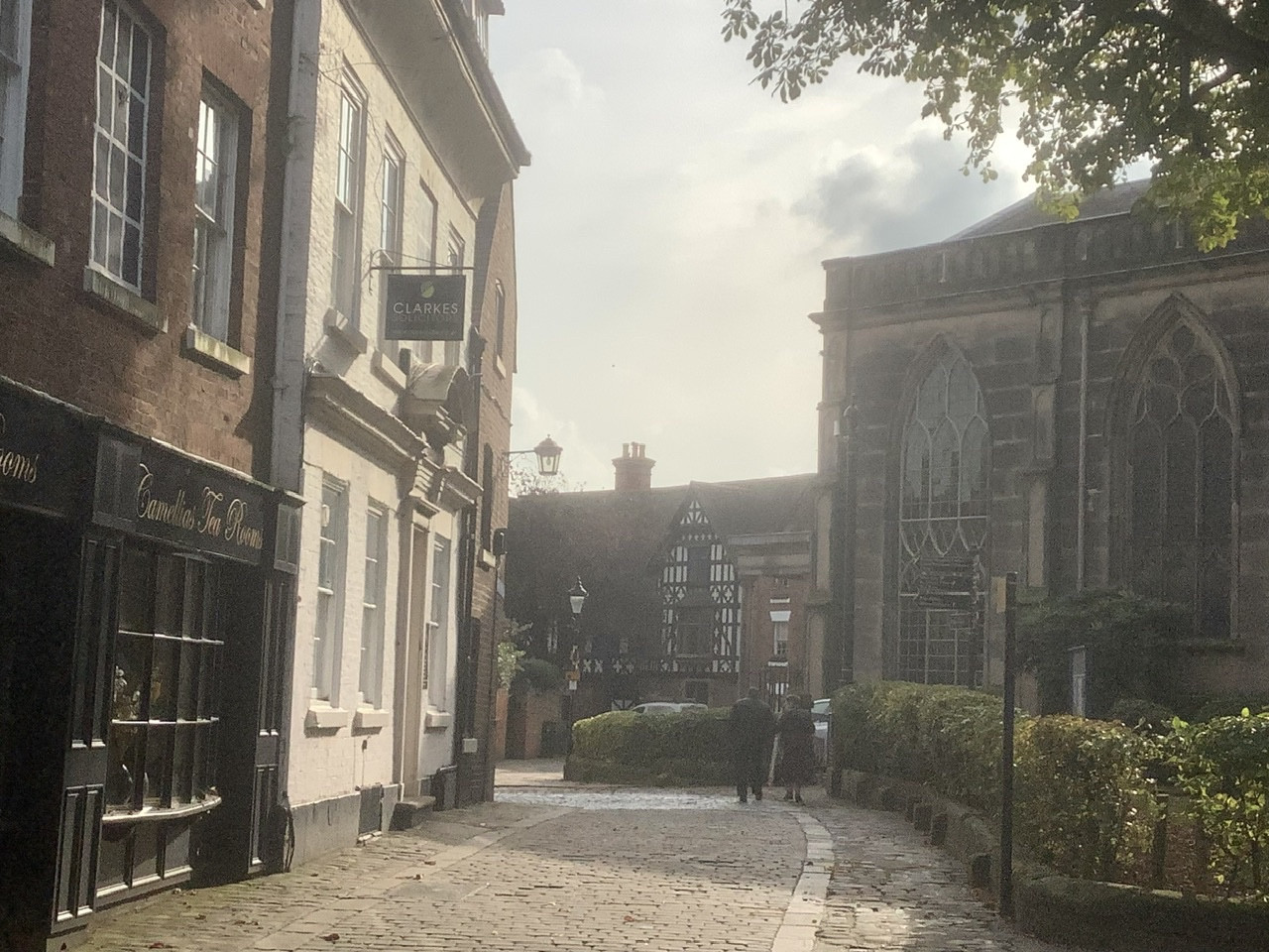 A winding, narrrow street with old buildings leaning in over it on the left hand side. There is a gothic looking church on the right hand side and two elderly people walking towards the end of the street. The light is hazy and warm.