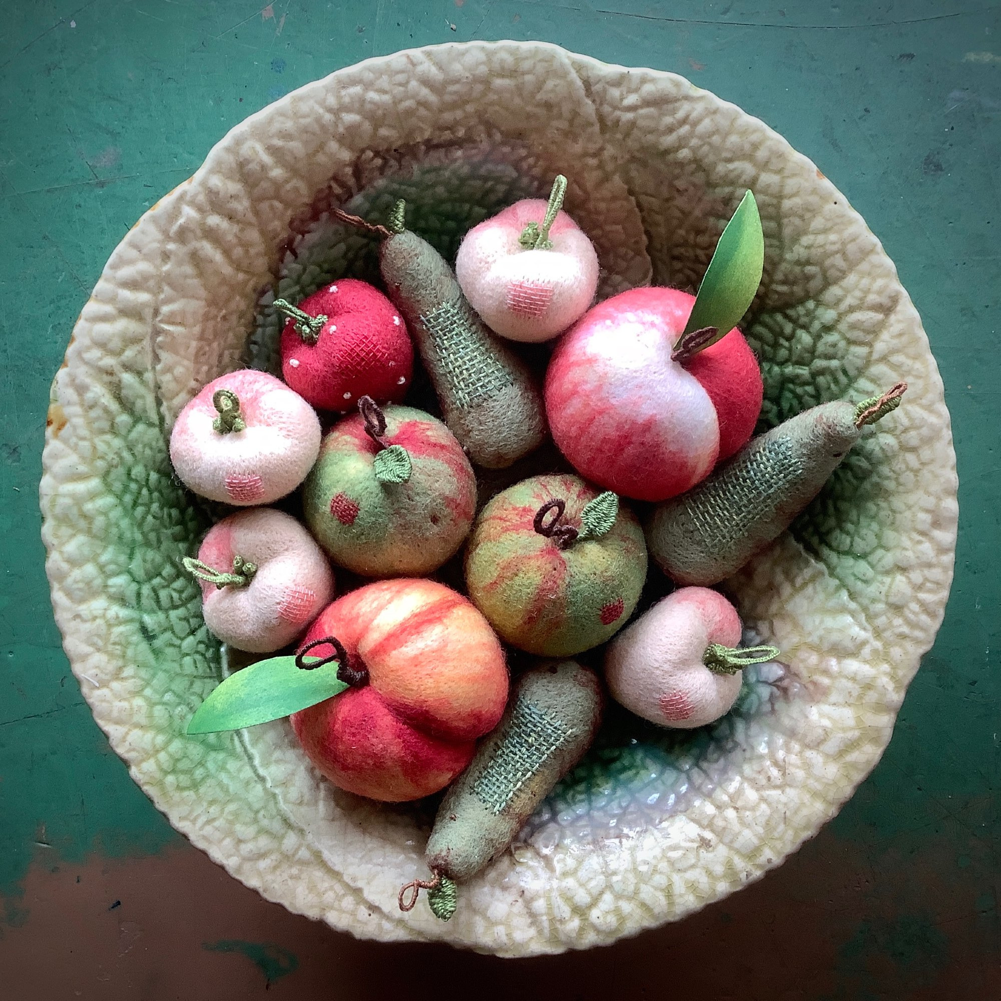 An assortment of woollen fruit baubles, long pears, round apples and peaches and small knobbly strawberries, all arranged on an old flat dish with a wrinkled texture like a cabbage.