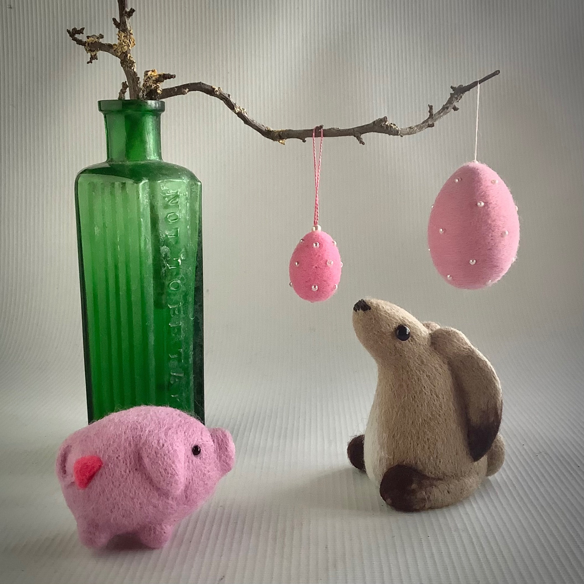 An old green straight sided bottle which has a small, lichen covered twig sticking out of the top. Hanging from the twig are two small pink eggs with pearl beads  studding them. In front of the vase is a pink woollen pig and to the right of the pig is a woollen model of a sitting baby hare with its ears laid back.