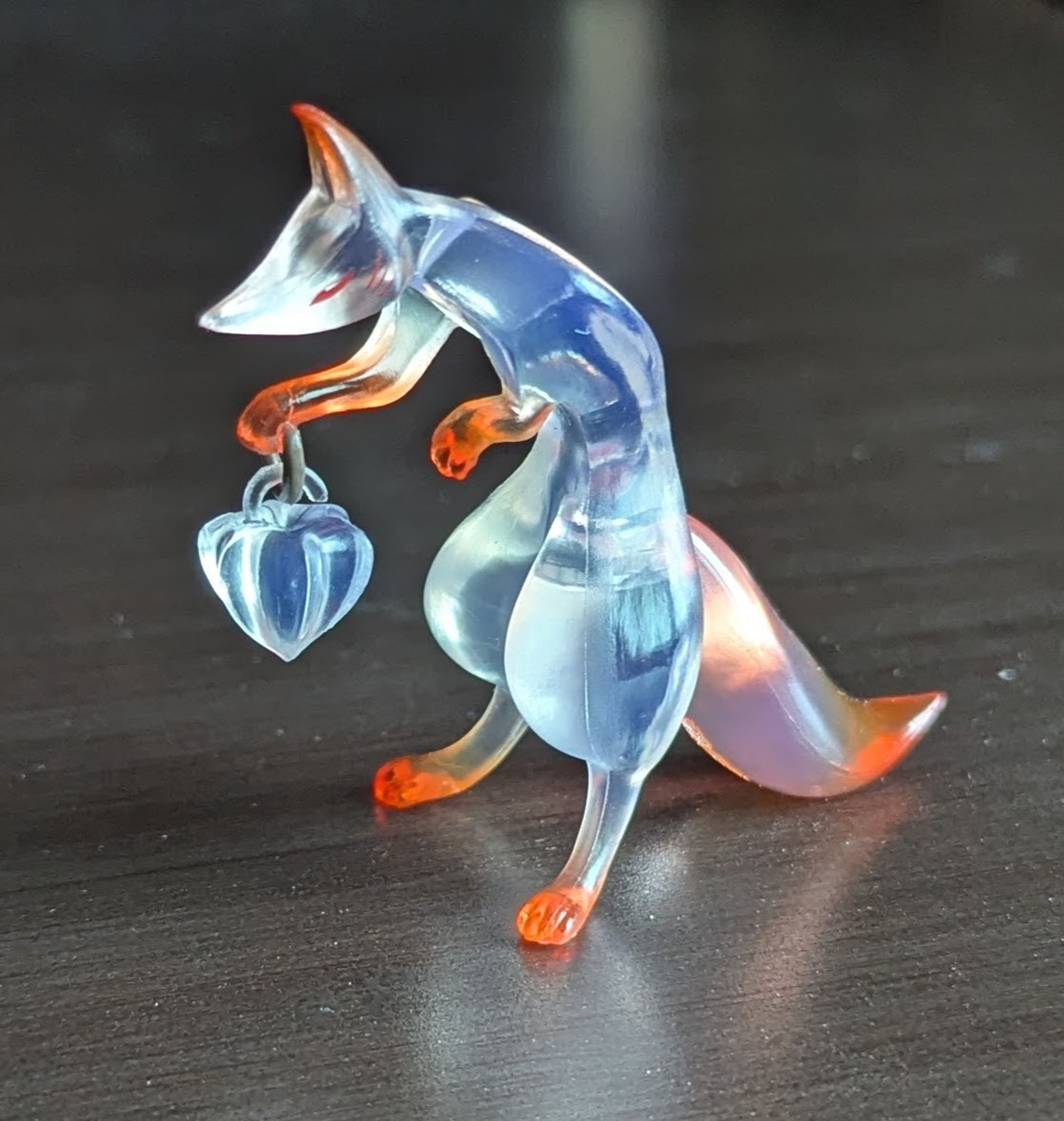 A clear fox figurine with orange-tinted ears, paws, and tail. It's holding a lantern shaped like a hōzuki (Scientific Name: Alkekengi officinarum).

This nightshade plant is also known as the Japanese Lantern or Chinese Lantern because its fruit is encased in a bright orange/red, papery pod that looks like a paper lantern. They're given as offerings during Japan's Bon festival to guide the souls of the dead.

Manufacturer's name for figurine: Cyber Carpenter Tōko