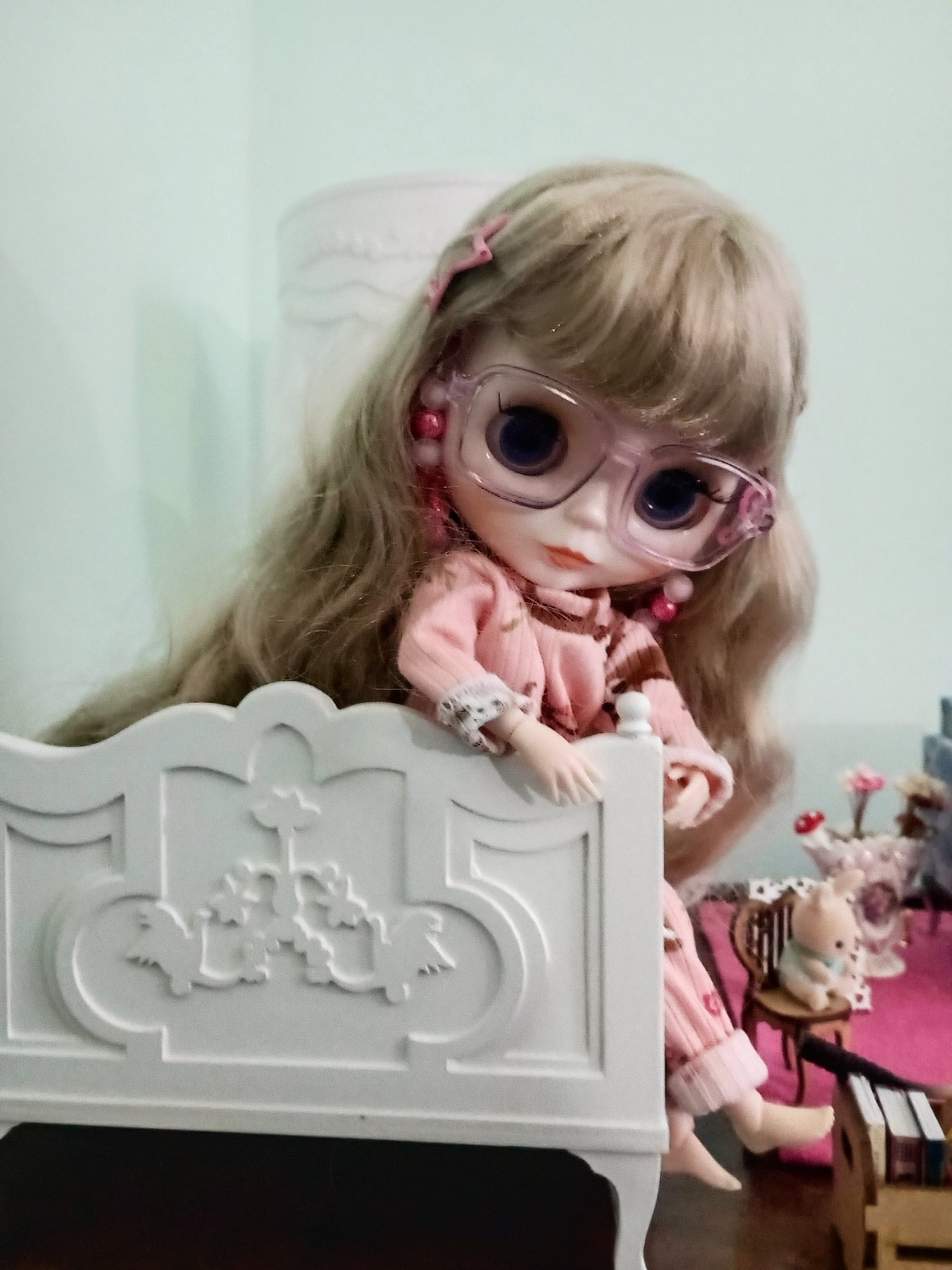 Boneca Blythe de pijama rosa sentada na beira de uma cama branca do tamanho dela.