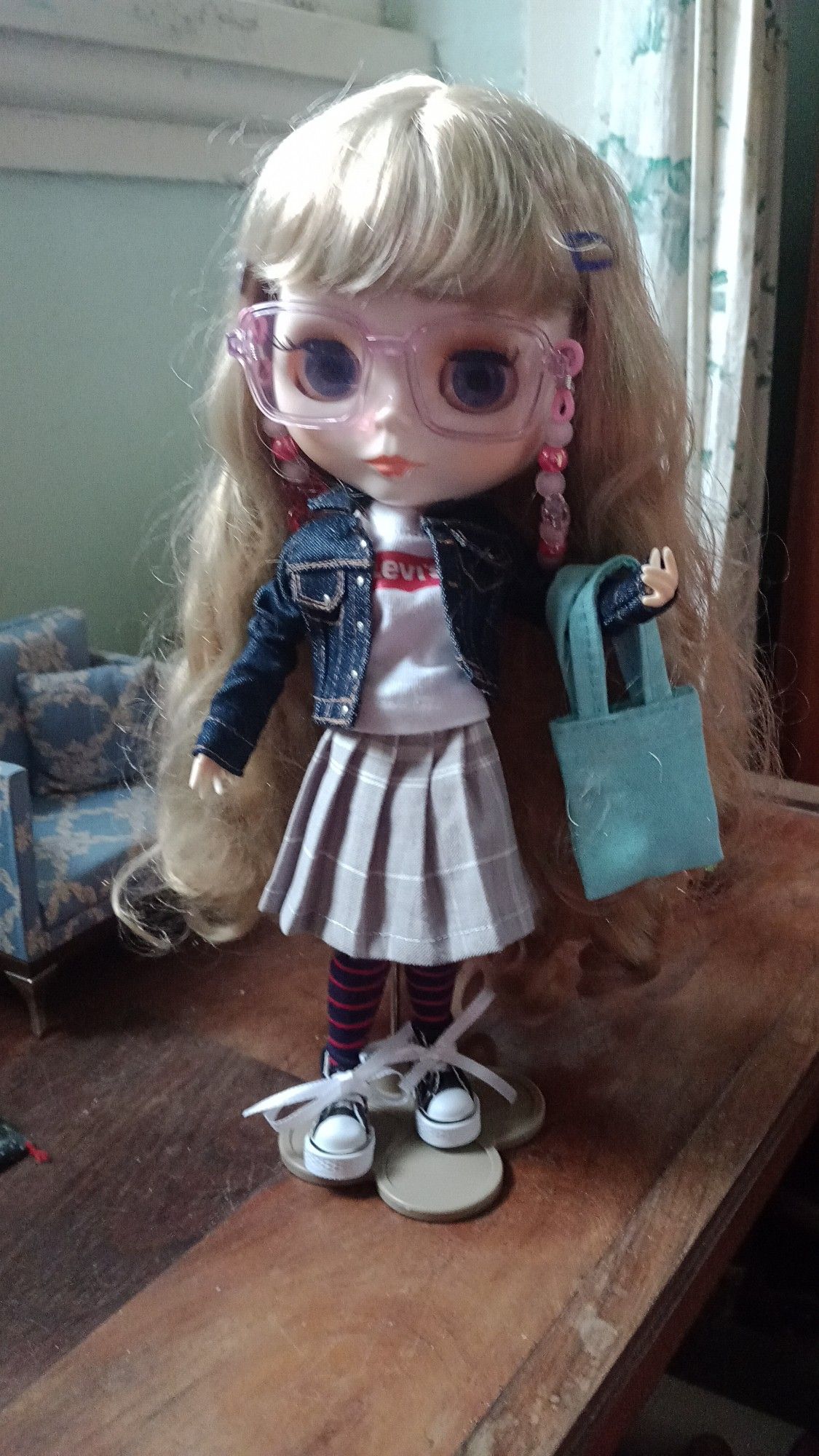 Boneca Blythe de óculos, saia de pregas, camiseta e jaqueta jeans, usando meias e um all star preto. Ela está carregando uma ecobag azul.