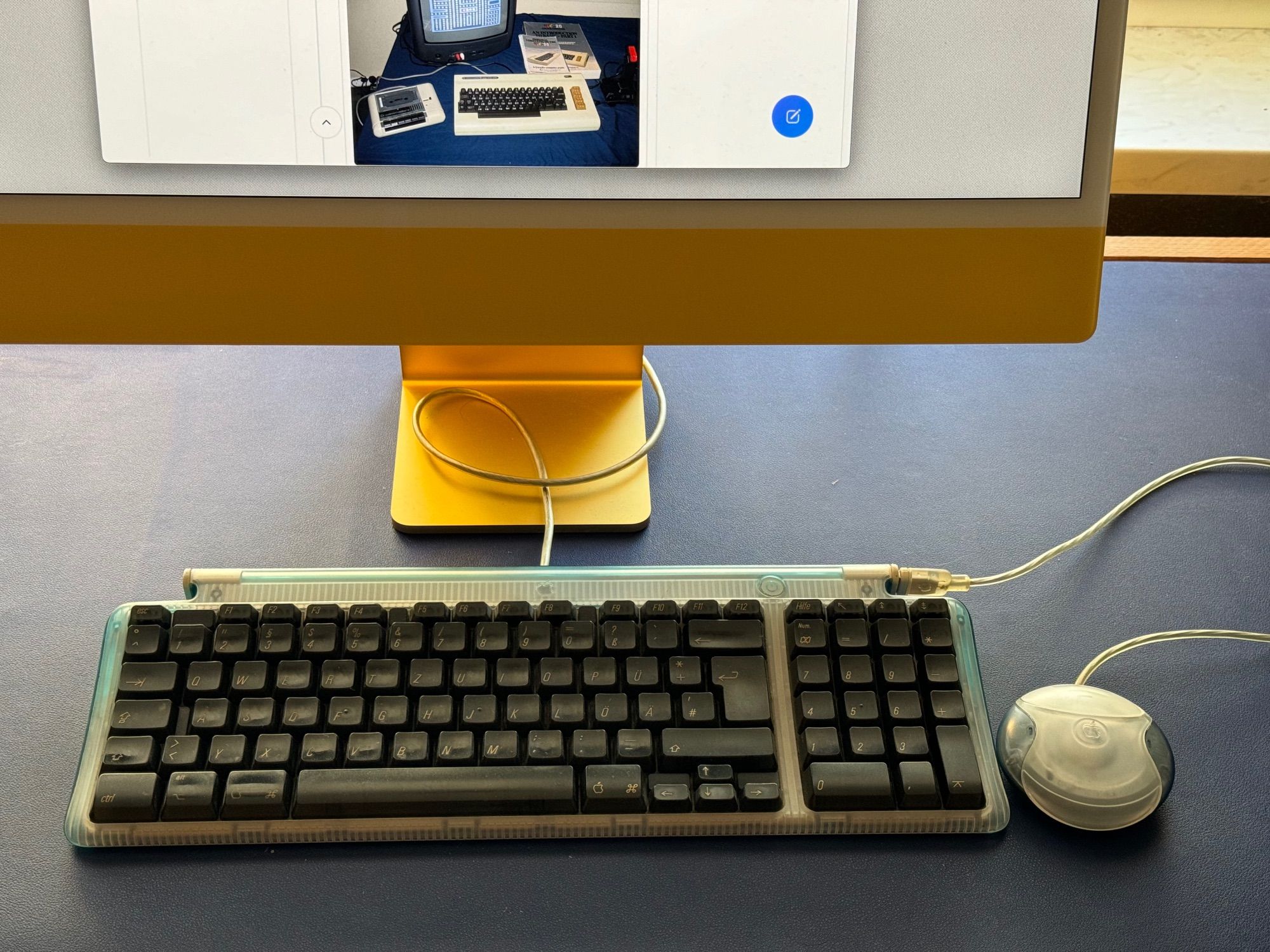 A Bondi blue keyboard from a German iMac G3 next to a grey puck mouse. All connected to a yellow iMac M1.