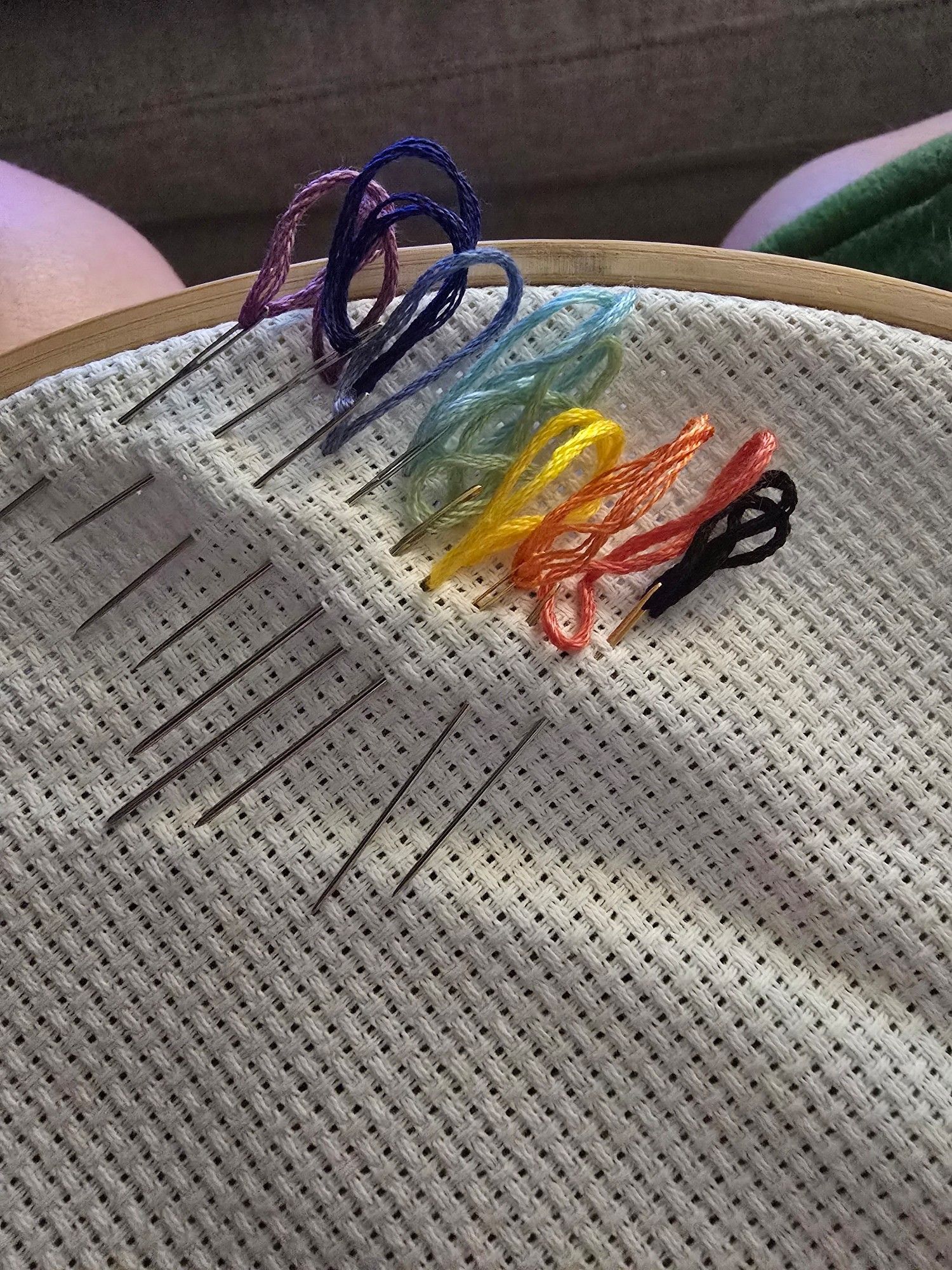 A white cross stitch canvas in a wooden loop with nine rainbow threads and needles set up