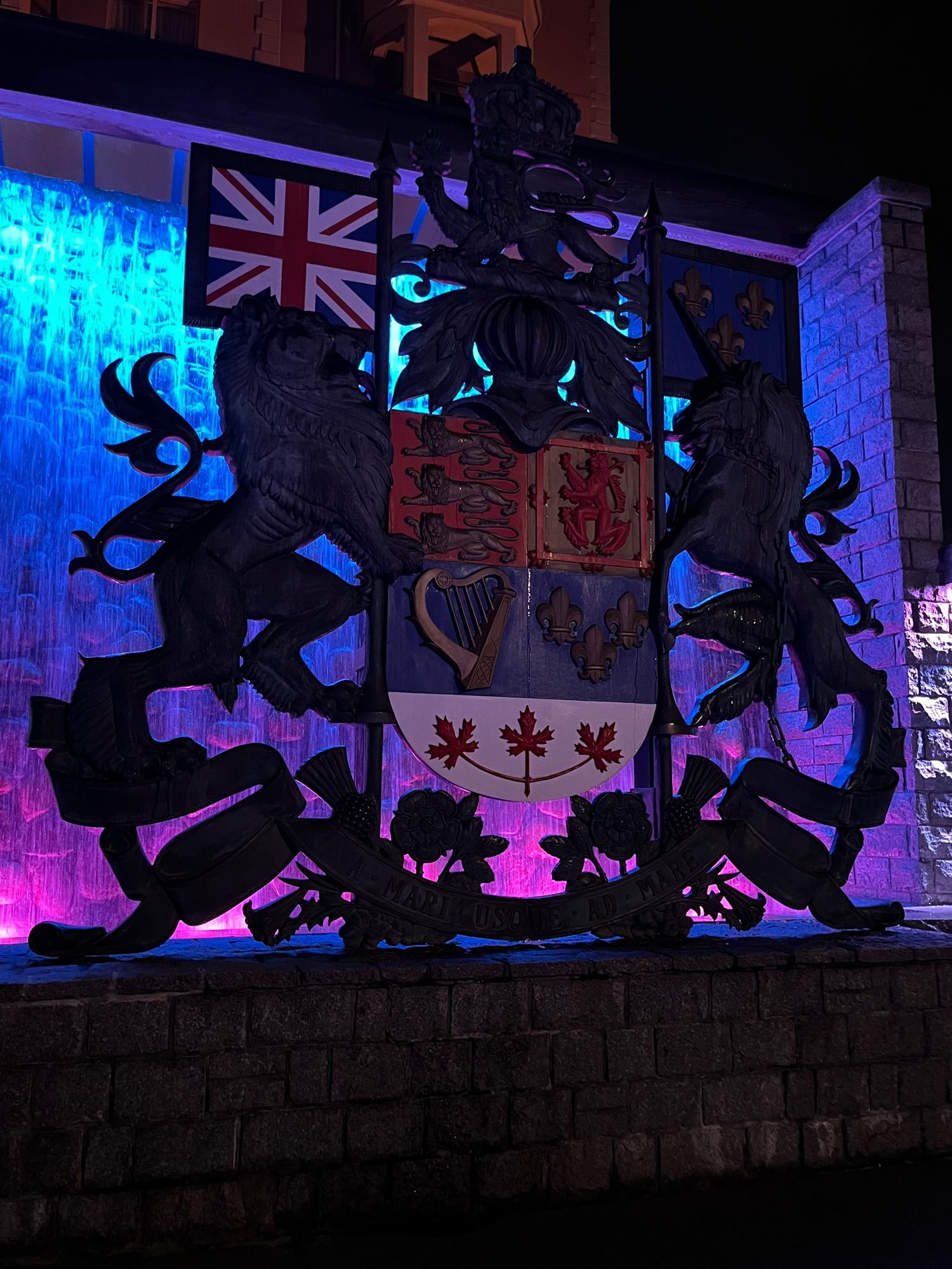 sculpture of canada's crest. 

Upon a royal helmet, a lion passant guardant or imperially crowned proper and holding in the dexter paw a maple leaf gules.

Tierced in fess, the first and second divisions containing the quarterly coat following, namely, 1st gules three lions passant guardant in pale or, 2nd, or a lion rampant within a double tressure flory-counter-flory gules, 3rd, azure a harp or stringed argent, 4th, azure three fleurs-de-lis or, and the third division being argent three maple leaves conjoined on one stem proper.

Dexter: a lion or holding a lance argent, point or, flying therefrom to the dexter the Royal Union Flag, sinister: a unicorn argent armed, crined and unguled or, gorged with a coronet composed of crosses-patée and fleurs-de-lis a chain affixed thereto and reflexed or, holding a like lance flying therefrom to the sinister a banner azure charged with three fleurs-de-lis or.

A wreath of roses, thistles, shamrocks, and lilies proper

A Mari usque ad Mare