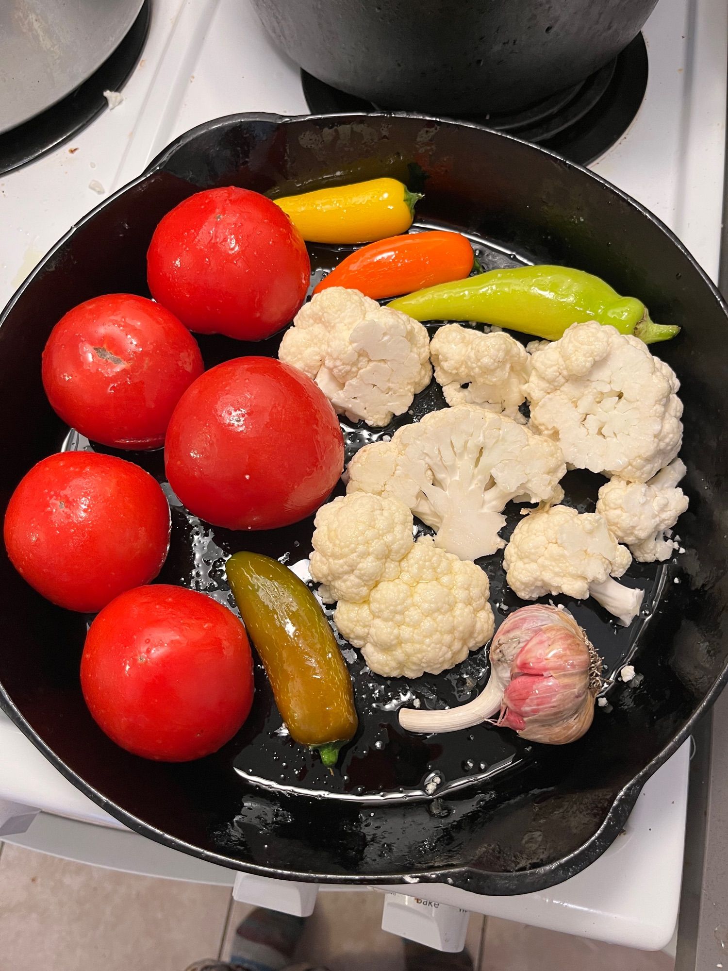 a cast iron pan with some small hot peppers, a head of garlic, cauliflower and five big red tomatoes