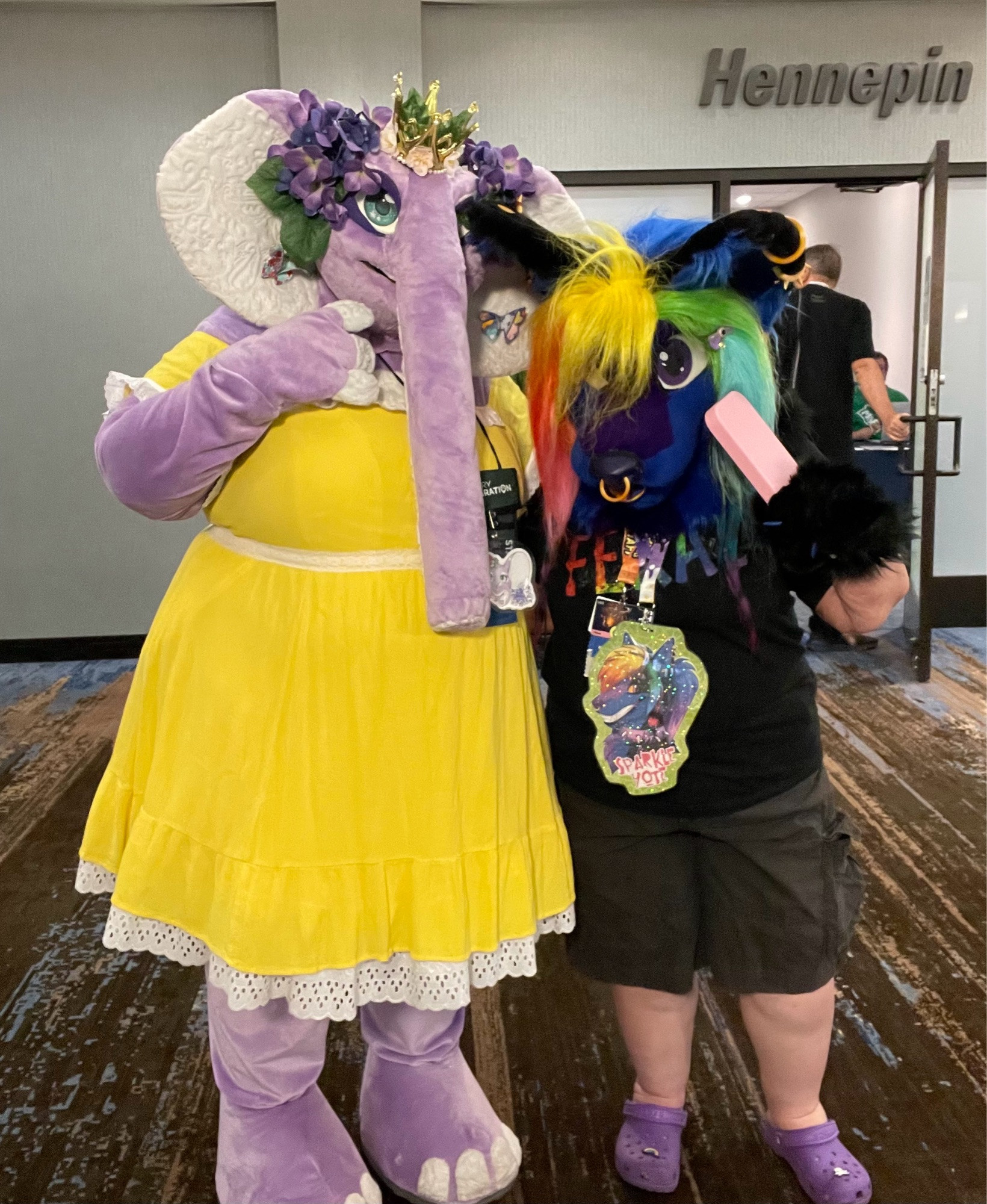 Lavender the elephant wearing a bright yellow dress and a crown, posing with Sparkle Yote, who has an ice cream shaped fan