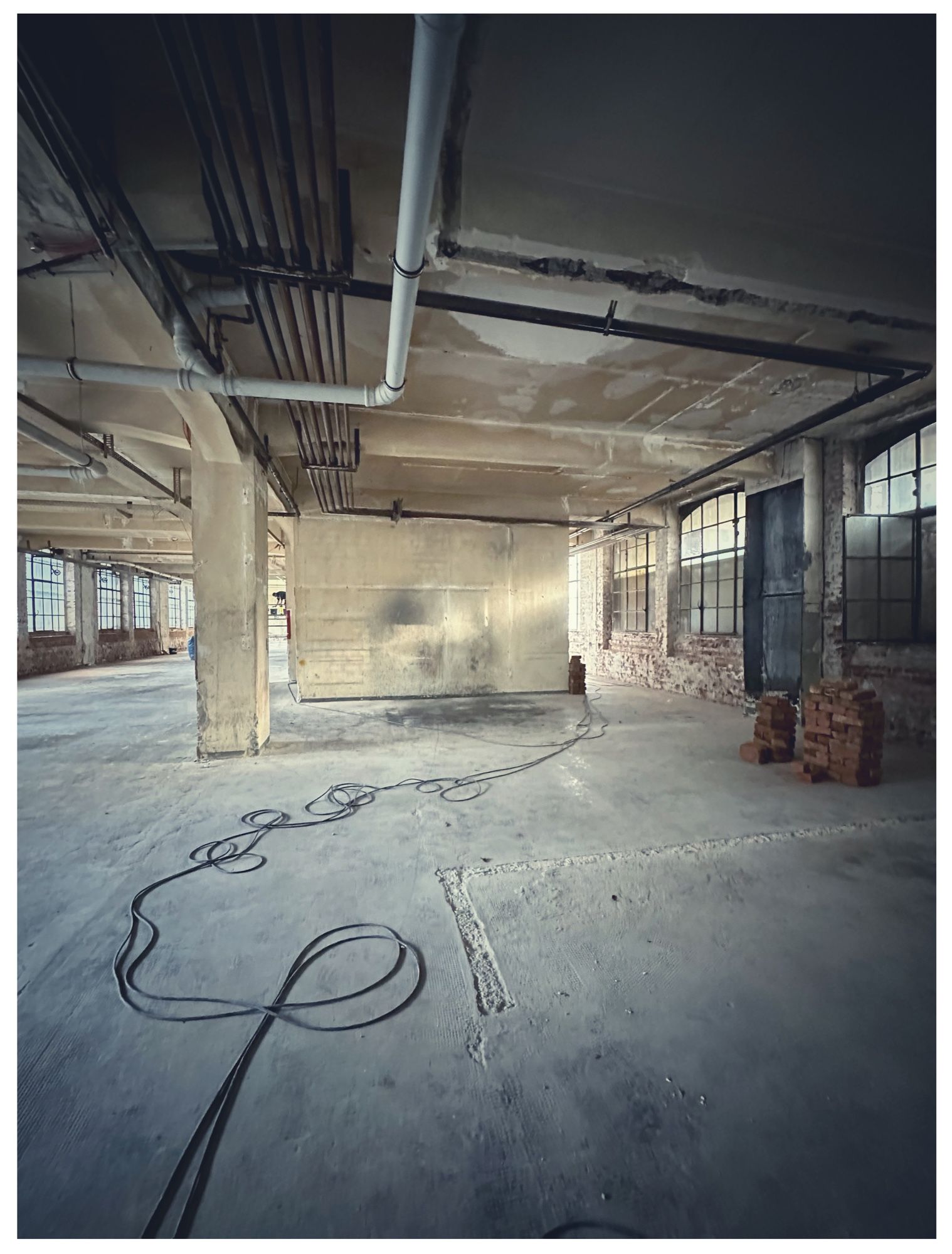 Leerstehende Industriehalle mit Fenstern rechts und links, Betonstützpfeiler und in der Flucht eine Trennmauer in der Mitte. Ein doppeltes Kabel schlängelt sich kunstvoll durch das ganze Bild.